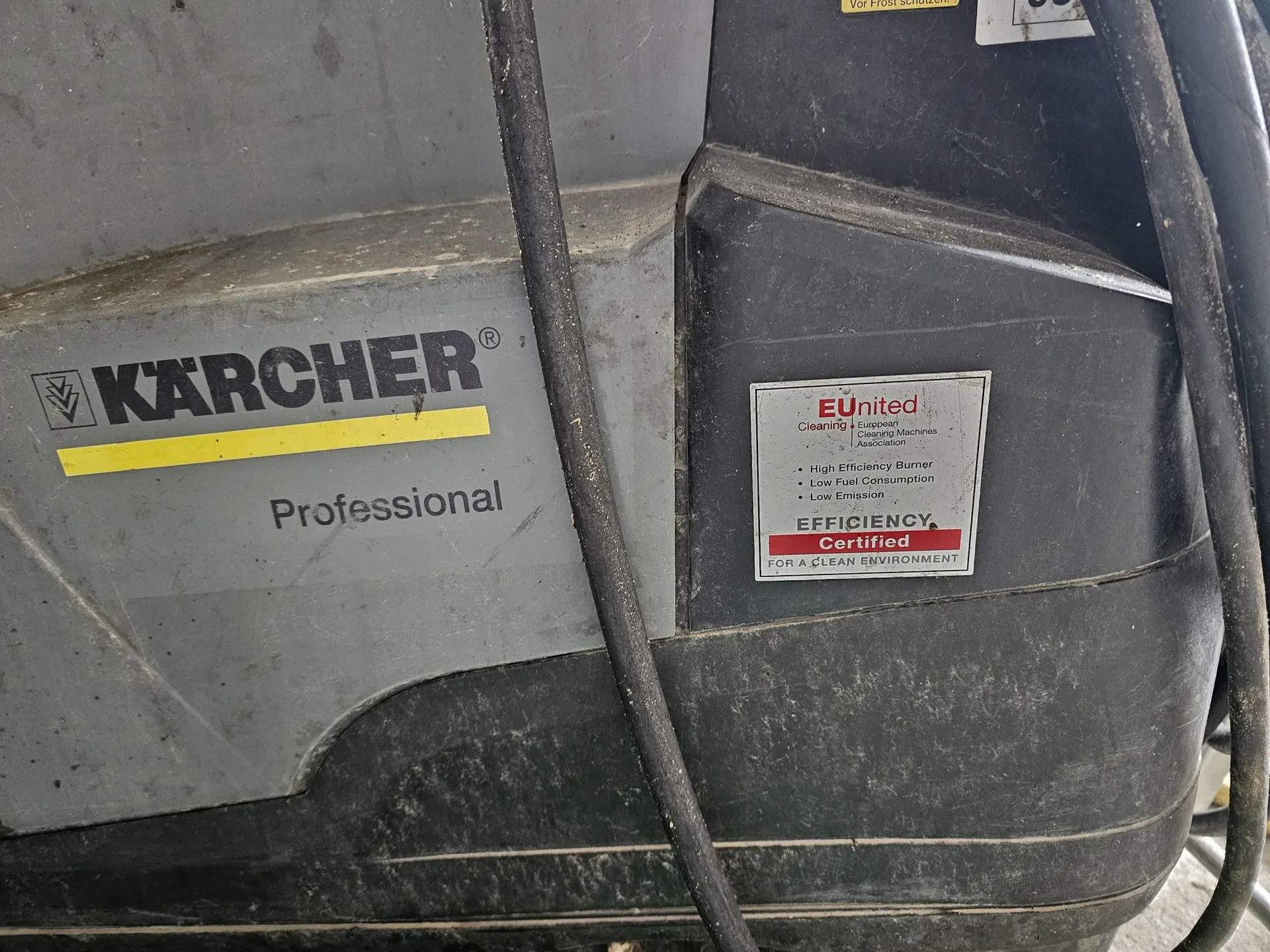Zbliżenie na szary, plastikowy korpus urządzenia marki Kärcher. Po lewej stronie widnieje logo 'KÄRCHER' z żółtym paskiem poniżej oraz napis 'Professional'. Po prawej stronie znajduje się naklejka z napisem 'EUunited Cleaning Equipment Cleaning Machines Association', listą cech: 'High Efficiency Burner', 'Low Fuel Consumption', 'Low Emission' oraz dużym napisem 'EFFICIENCY Certified FOR A CLEAN ENVIRONMENT'. Przez środek obrazu przebiegają dwa czarne kable elektryczne.