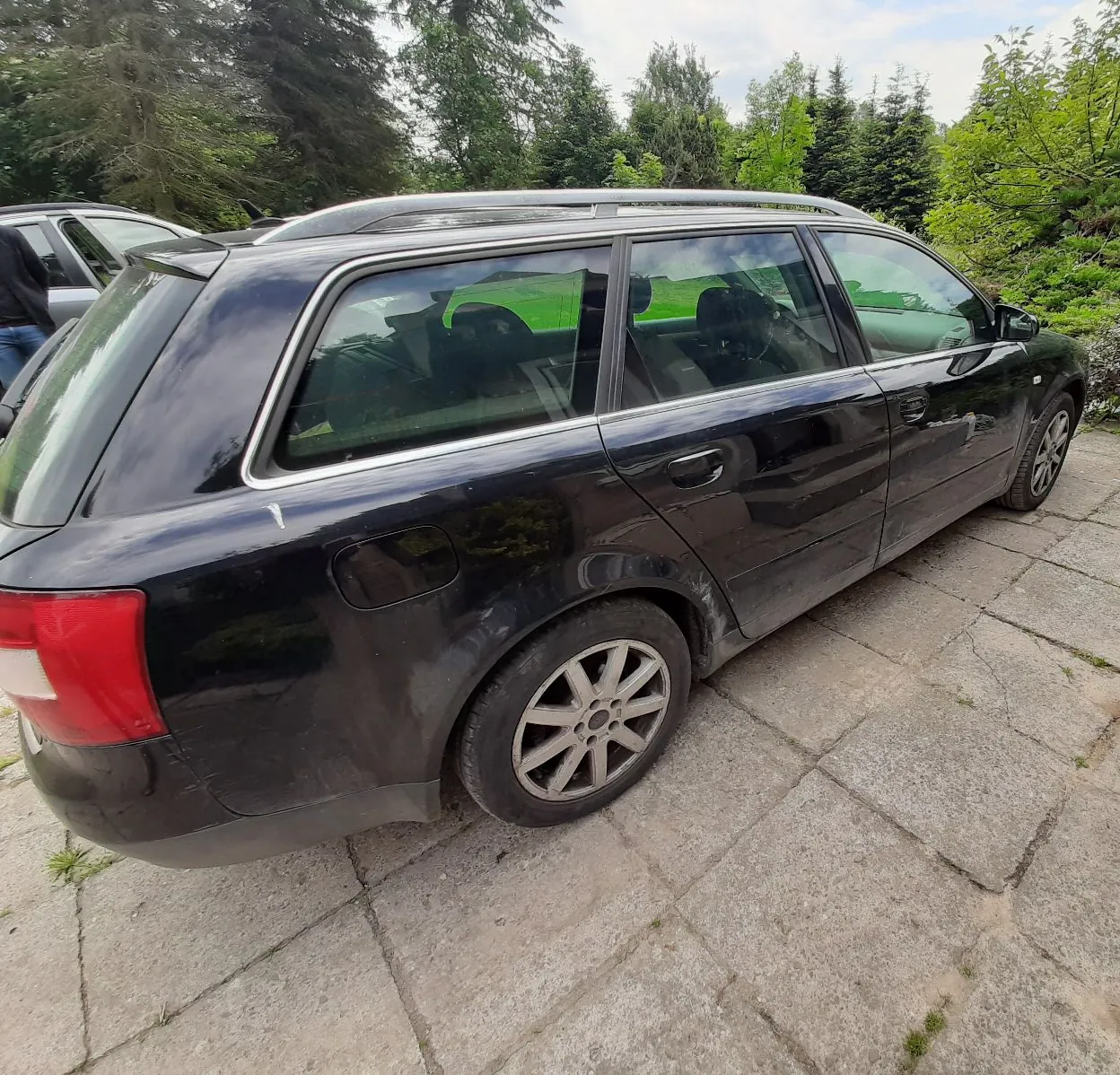 Czarny samochód typu kombi zaparkowany na betonowym podjeździe. Pojazd jest widoczny z profilu od strony pasażera, z tyłu widać czerwone światło stopu. W tle znajdują się zielone drzewa i krzewy pod jasnym niebem.
