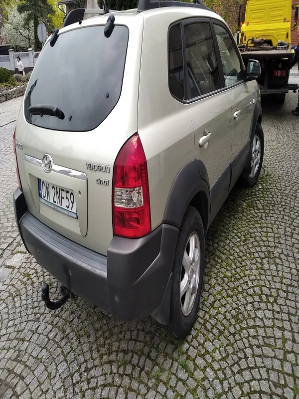 Zdjęcie przedstawia tył beżowego SUV-a marki Nissan z widocznym numerem rejestracyjnym DW 2NF59. Samochód stoi na brukowanej powierzchni z mchem między kamieniami. W tle widoczny jest żółty pojazd ciężarowy oraz fragment ogrodzenia i drzew. Okna samochodu są przyciemnione, a na tylnym zderzaku znajduje się hak holowniczy.