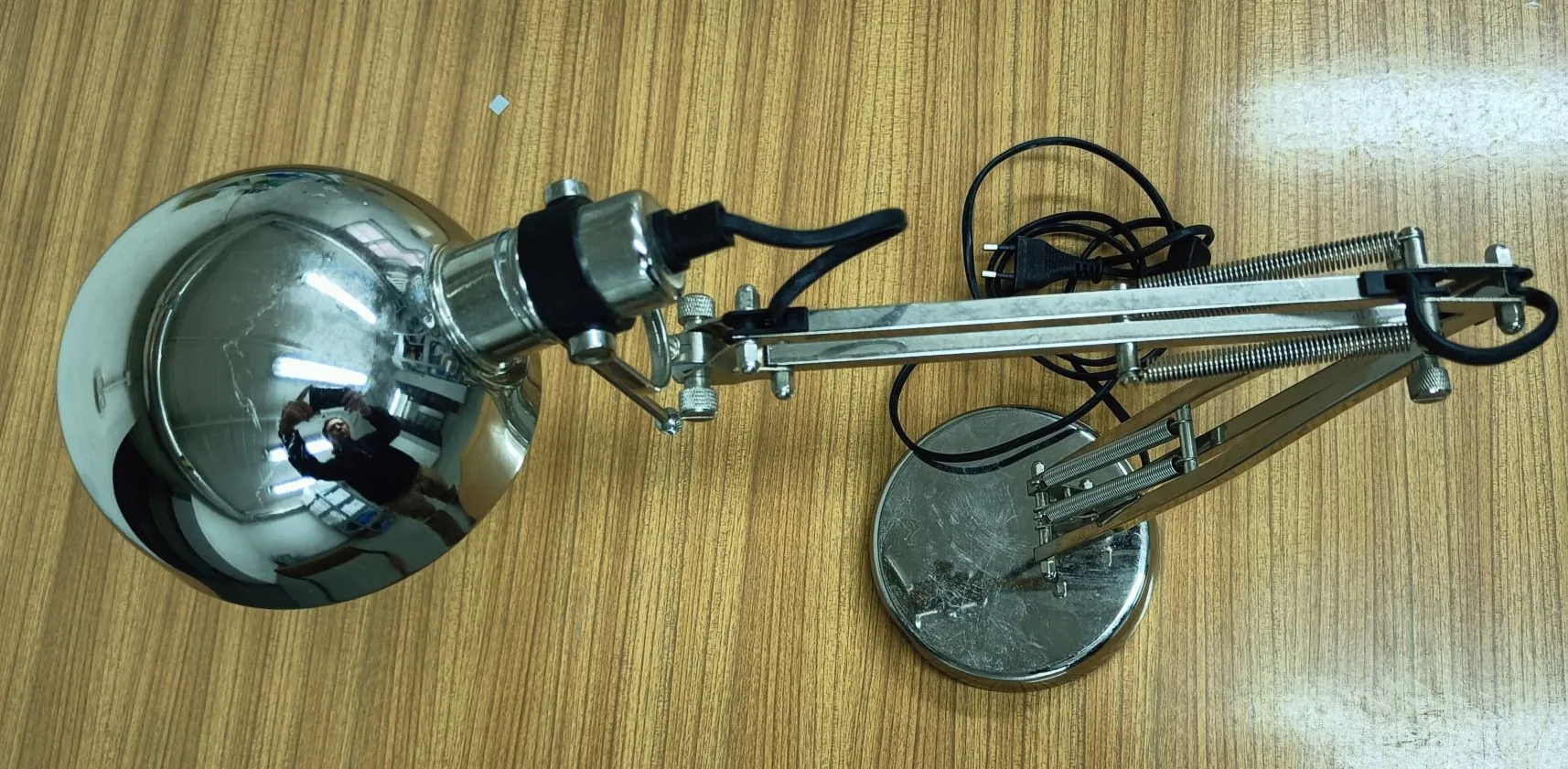 Srebrna lampka biurkowa z metalowym, ruchomym ramieniem i okrągłym kloszem, leżąca na drewnianym blacie. Lampka ma czarny kabel zasilający i okrągłą podstawę.