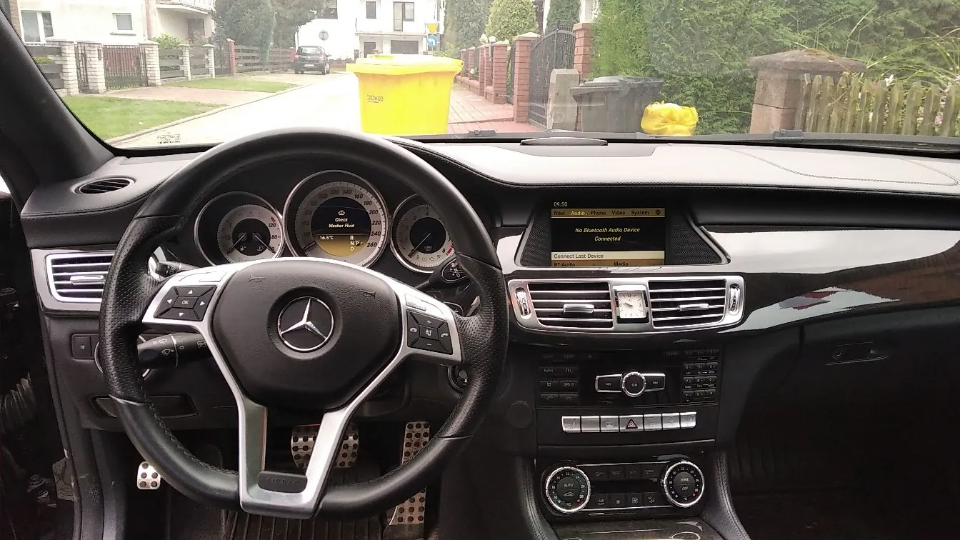 Widok z wnętrza samochodu marki Mercedes-Benz z perspektywy kierowcy. Widoczna jest czarna skórzana kierownica z logo marki w centrum, zestaw wskaźników z prędkościomierzem i obrotomierzem oraz centralny ekran multimedialny z napisem 'No Bluetooth Audio Device Connected'. Poniżej ekranu znajdują się przyciski sterowania radiem i klimatyzacją. Przez przednią szybę widoczna jest ulica z domami, żółtym kontenerem na odpady oraz zielenią.
