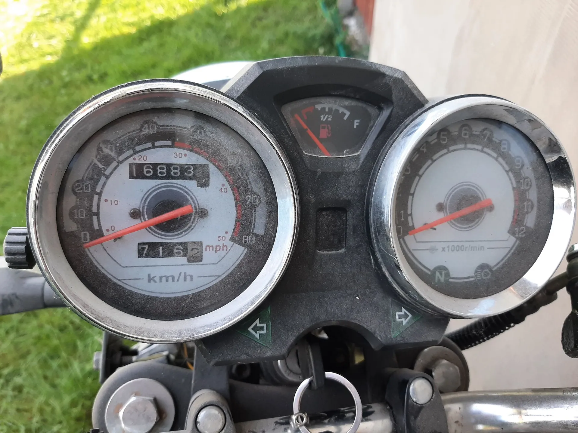 Zbliżenie na zestaw wskaźników motocyklowych. Po lewej znajduje się prędkościomierz z tarczą w km/h i mph, z licznikiem przebiegu wskazującym 16883 oraz mniejszym licznikiem 716. W centrum znajduje się wskaźnik poziomu paliwa. Po prawej znajduje się obrotomierz z oznaczeniem x1000r/min. Poniżej wskaźników widoczne są kontrolki kierunkowskazów oraz stacyjka z kluczykiem.