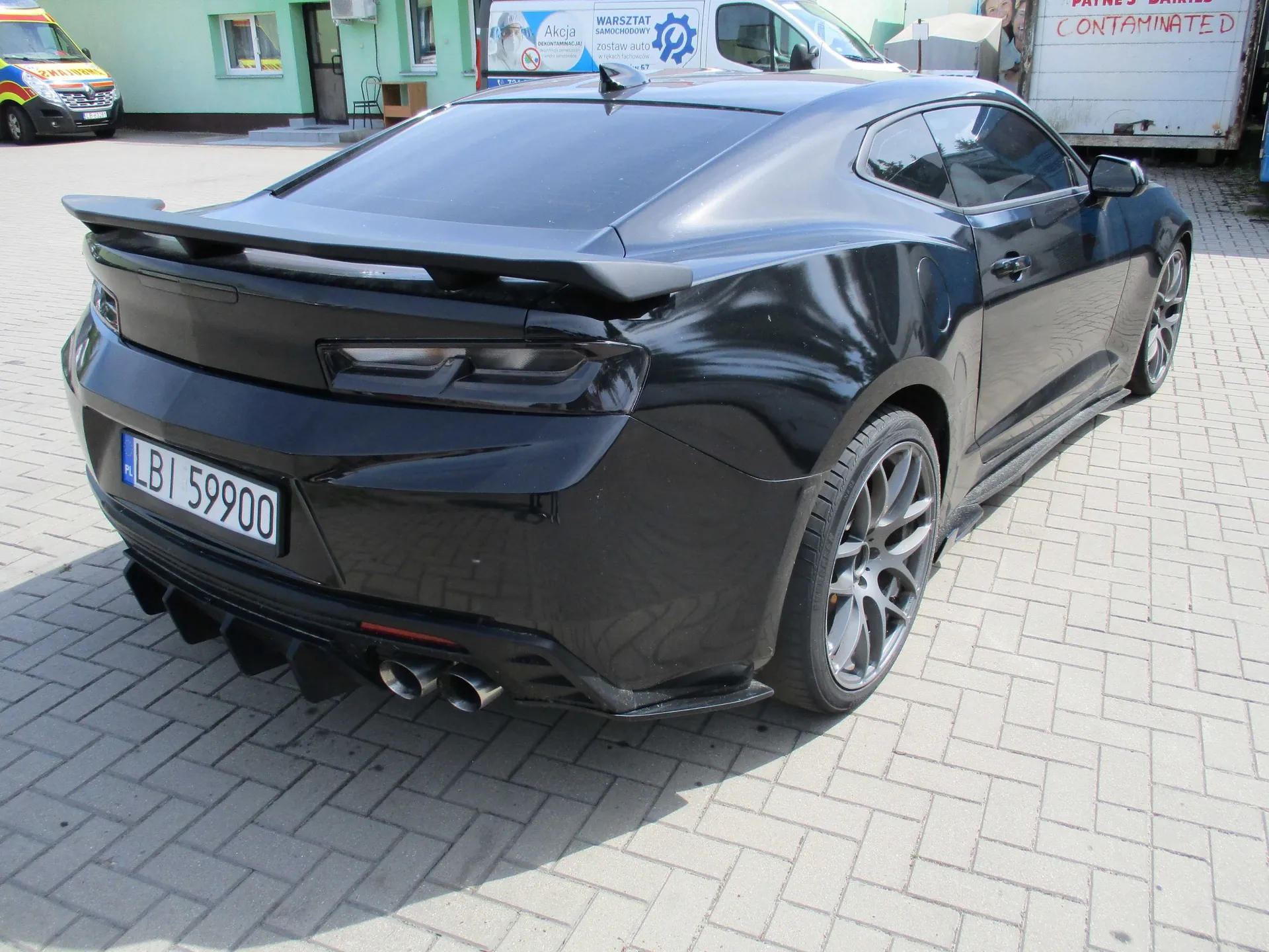 Czarny samochód sportowy typu coupe, widoczny od tyłu pod kątem. Pojazd posiada spojler na klapie bagażnika, dyfuzor z dolnym profilem oraz podwójne wydechy z każdej strony. Tablica rejestracyjna to LBI 59900. Samochód stoi na kostce brukowej przed budynkiem z szyldem 'WARSZTAT SAMOCHODOWY'. W tle widoczny jest fragment żółtego vana oraz biała przyczepa z napisem 'CONTAMINATED'.