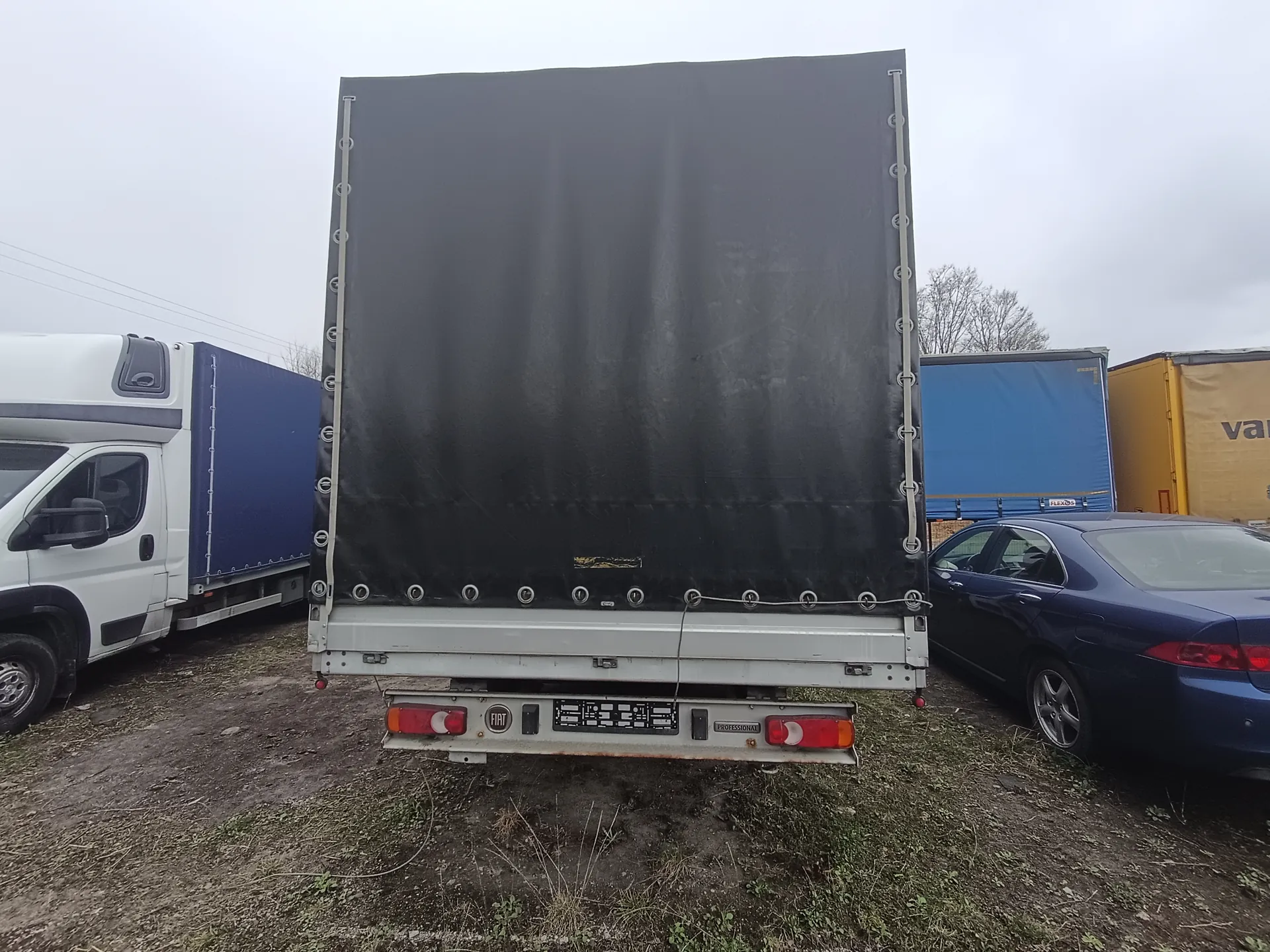 Tył ciężarówki z czarnym namiotem, białym zderzakiem i tablicą rejestracyjną. Widoczne logo 'FIAT' oraz napis 'var' na żółtym pojeździe w tle. Po lewej stronie biała furgonetka z niebieskim namiotem, po prawej niebieski samochód osobowy. Tło: niebo, drzewa i inne pojazdy. Podłoga z trawą i błotem.