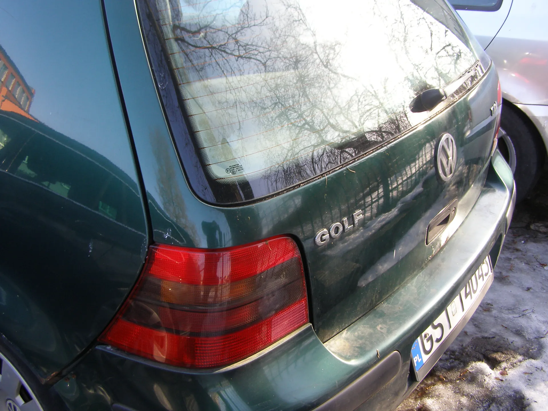 Zbliżenie na tył zielonego samochodu Volkswagen Golf. Widoczne są napisy 'GOLF' i logo VW na bagażniku, czerwone światło tylnego reflektora oraz tablica rejestracyjna z polskim kodem 'PL' i numerem 'GS 14049'. W tle odbicie drzew bez liści w szybie, fragment innego pojazdu oraz śnieg na ziemi.
