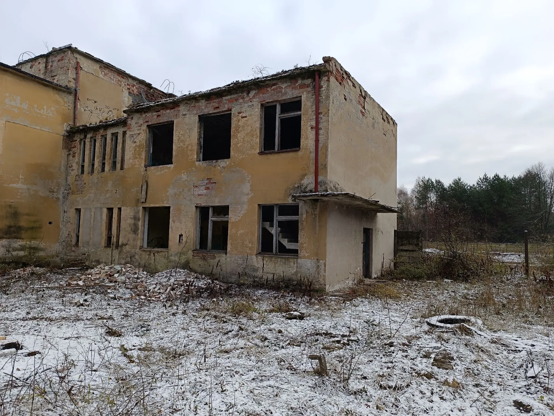 Opuszczony, dwupiętrowy budynek z odpadającym tynkiem w kolorze żółtym i beżowym. Budynek jest w stanie ruiny, brakuje szyb w większości okien, a dach jest częściowo zniszczony. Przed budynkiem znajduje się zarośnięty teren pokryty cienką warstwą śniegu i gruzem. W tle widoczny jest ciemny pas lasu pod zachmurzonym niebem.
