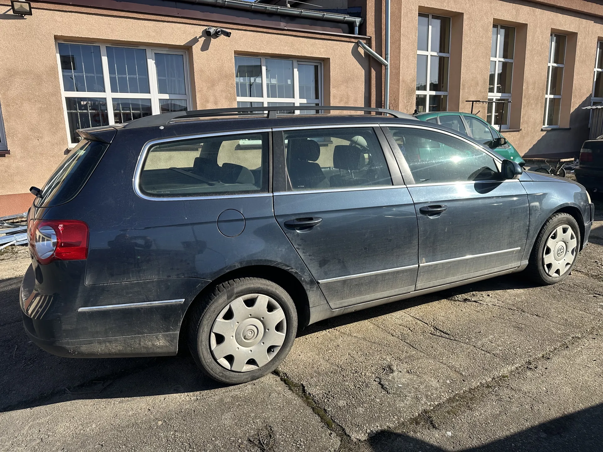 Ciemnoszary samochód typu kombi z dachowymi prętami, zaparkowany na betonowym placu przed beżowym budynkiem z białymi ramami okiennymi. W tle widoczny zielony samochód oraz fragment czarnego pojazdu. Na elewacji budynku znajdują się okna i kamera monitorująca. Betonowa powierzchnia posiada pęknięcia i ślady zużycia.