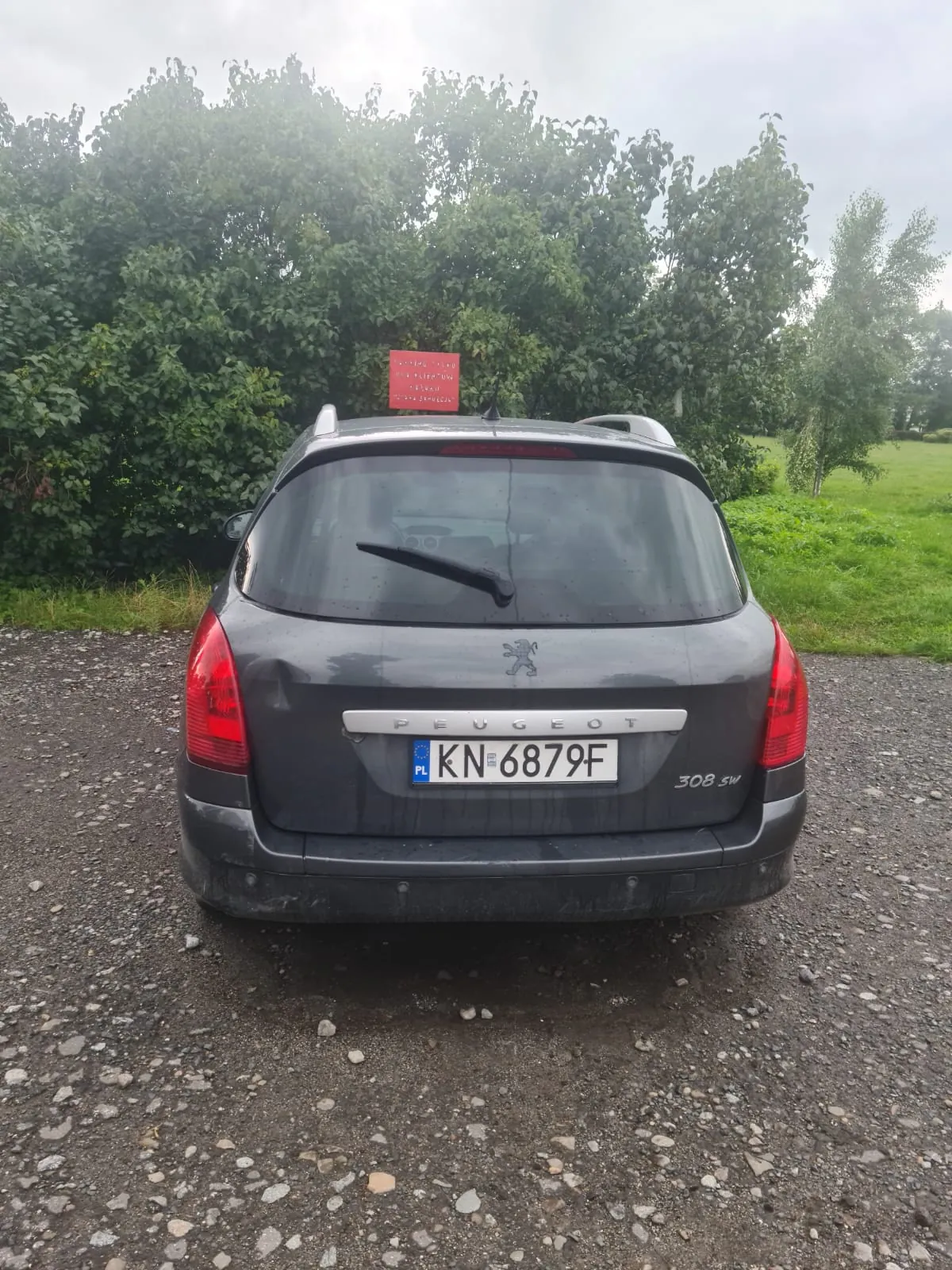Tylne widok ciemnoszarego samochodu Peugeot 308 SW zaparkowanego na żwirowej powierzchni. Widoczna biała tablica rejestracyjna z polskim kodem "PL" i numerem "KN 6879F", logo marki oraz napis "308 SW". W tle zielone drzewa, trawa i czerwony znak z nieczytelnym tekstem. Niebo jest zachmurzone.