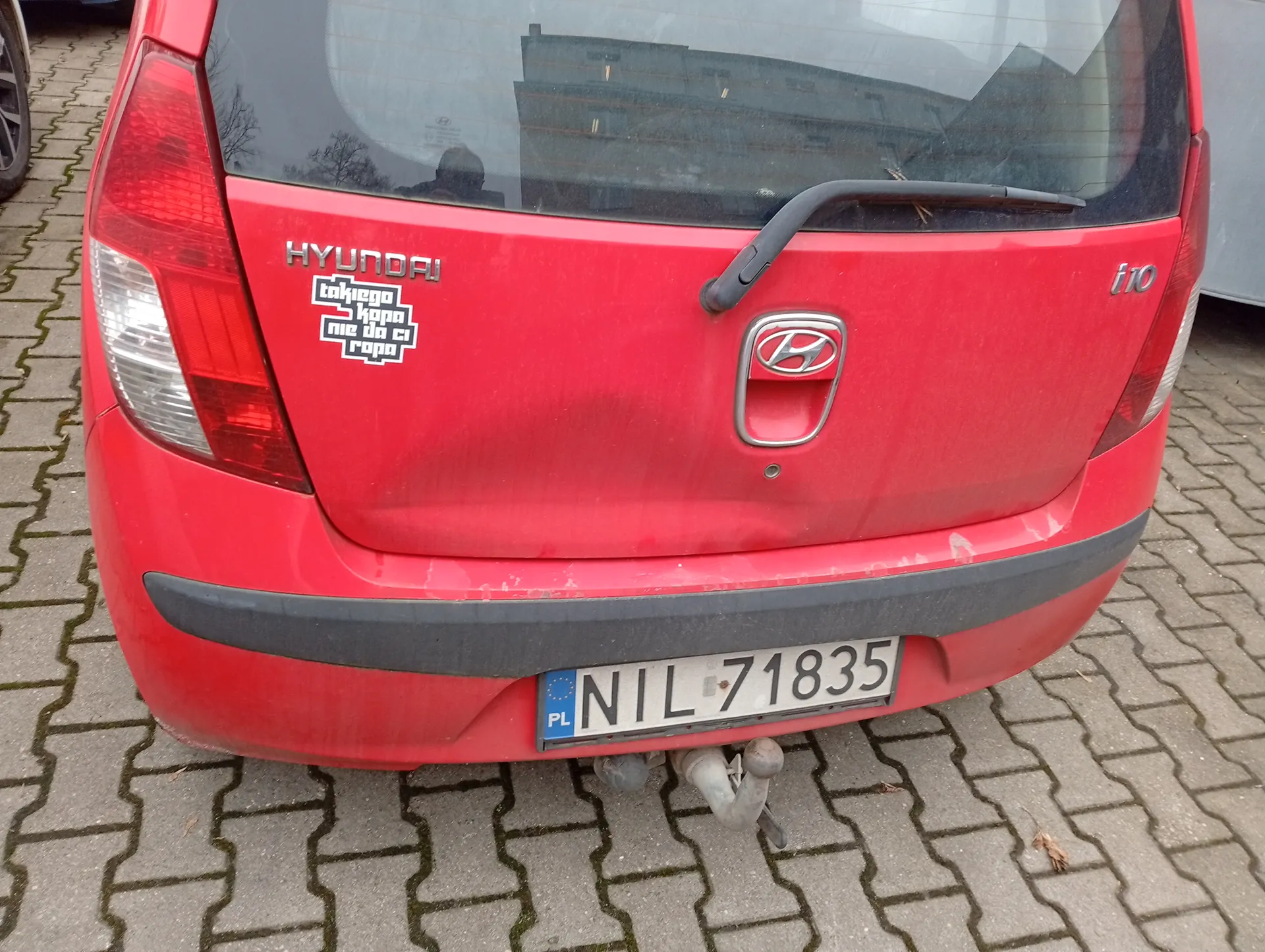 Czerwony samochód Hyundai i10 z tyłu, zaparkowany na brukowanej powierzchni. Widoczny jest biały numer rejestracyjny PL NIL 71835, logo marki w centrum, napis HYUNDAI po lewej stronie oraz model i10 po prawej. Na lewej stronie bagażnika naklejka z napisem takiego kopa nie da ci rópa. W tle odbicie budynku i osoby w szybie. Samochód ma czerwone światła tylna i szary zderzak.