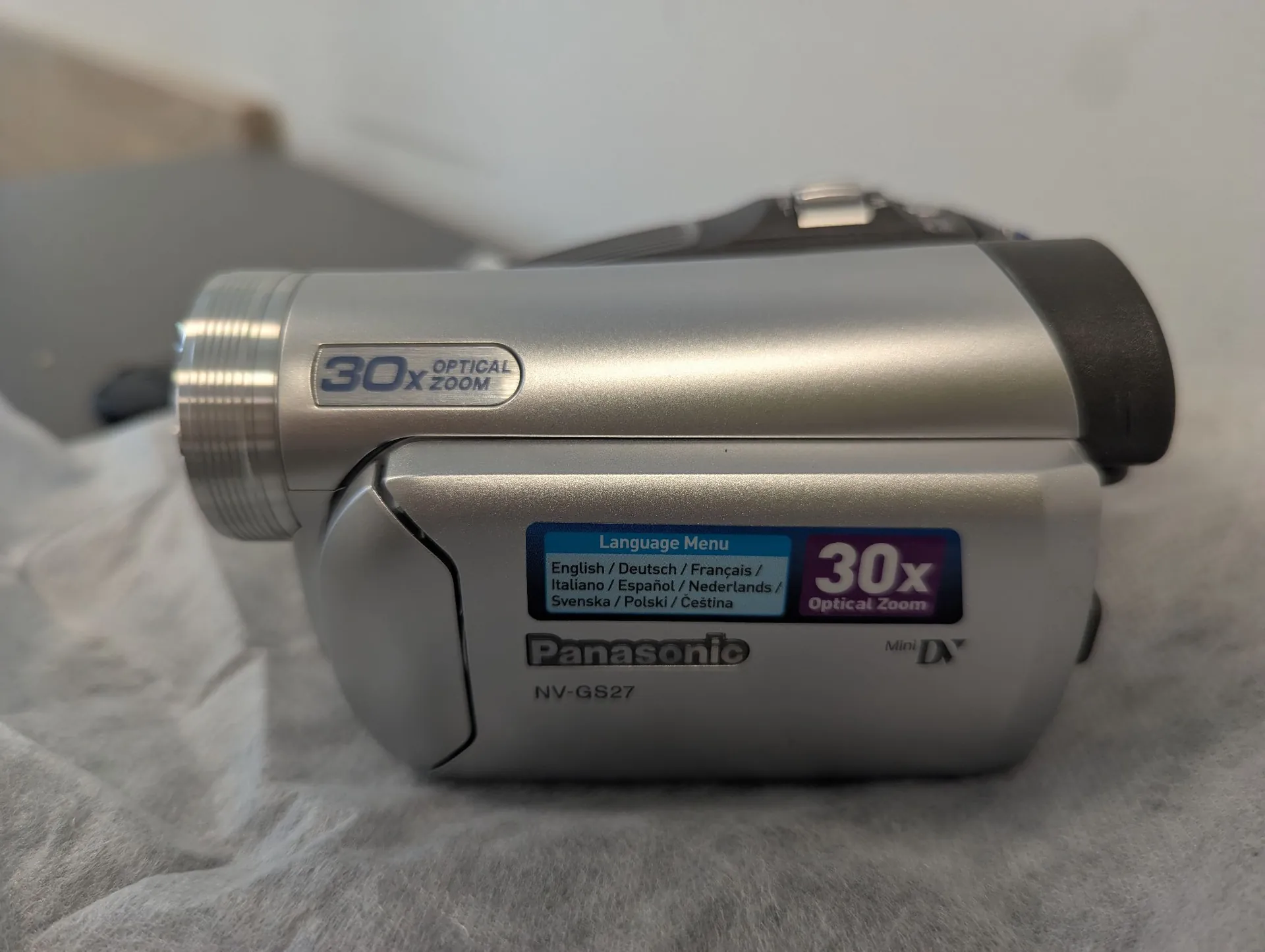 Srebrna kamera wideo marki Panasonic, model NV-GS27. Na obiektywie widnieje napis '30x OPTICAL ZOOM'. Na obudowie znajduje się naklejka 'Language Menu' z listą dostępnych języków, w tym polskiego, oraz napis '30x Optical Zoom' i logo 'Mini DV'.