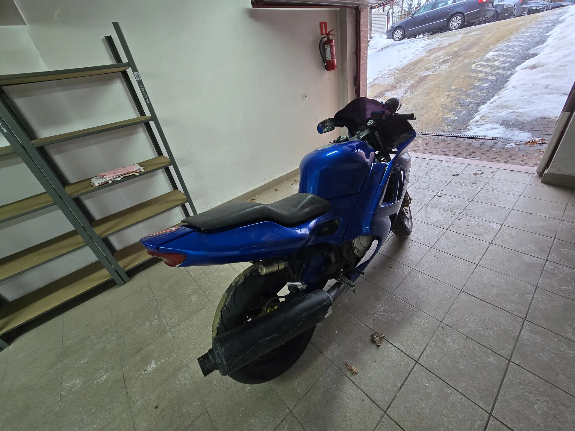Niebieski motocykl stojący w garażu z podłogą z kafli. Po lewej stronie metalowa półka z kilkoma papierami na jednej z półek. Na ścianie czerwony gaśnicz z białą cyfrą 1 na tabliczce powyżej. Za otwartymi drzwiami garażu widoczna zewnętrzna powierzchnia z resztkami śniegu, brukowana droga oraz zaparkowane samochody. Na podłodze w garażu kilka suchych liści.