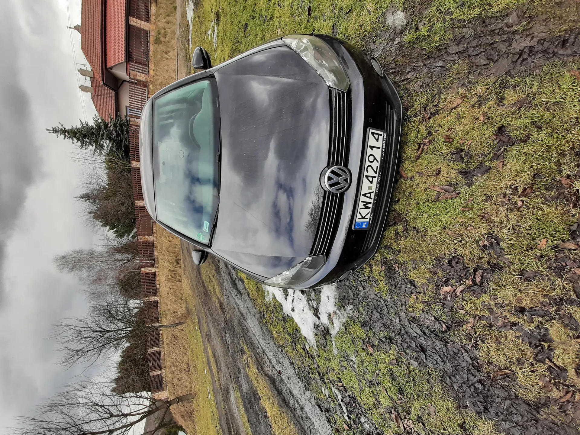 Czarny samochód marki Volkswagen z tablicą rejestracyjną KWA-42914 zaparkowany na trawiastym i błotnistym terenie. W tle dom z czerwonym dachem, drewniany płot, drzewa bez liści oraz pochmurne niebo. Na ziemi widoczne są ślady opon, resztki śniegu i sucha trawa.
