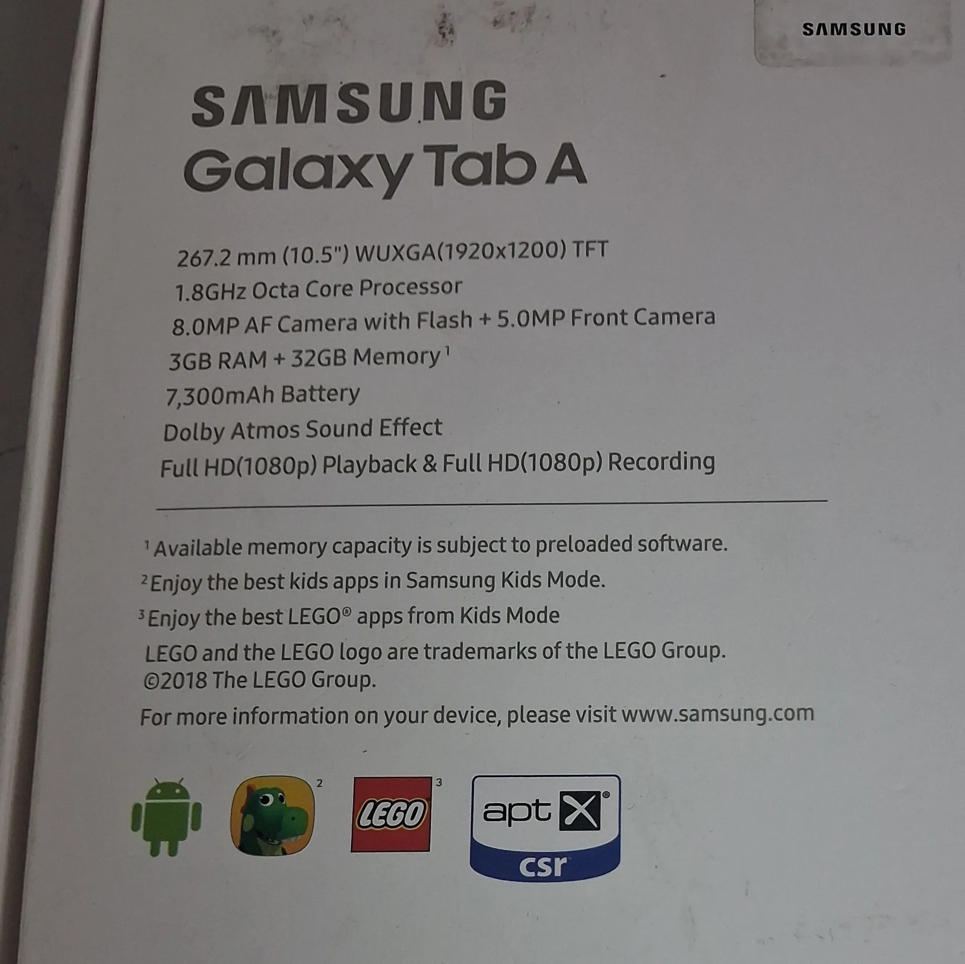 Zdjęcie tyłu białego pudełka tabletu Samsung Galaxy Tab A. Na opakowaniu znajduje się czarny tekst z nazwą urządzenia oraz specyfikacją techniczną: ekran 267.2 mm (10.5") WUXGA(1920x1200) TFT, procesor 1.8GHz Octa Core, aparat główny 8.0MP AF z lampą błyskową i przedni 5.0MP, 3GB RAM + 32GB pamięci, bateria 7,300mAh, efekt dźwiękowy Dolby Atmos oraz odtwarzanie i nagrywanie w Full HD(1080p). Poniżej znajdują się przypisy dotyczące pamięci i trybu Kids Mode z logo LEGO. Na dole widoczne są ikony: zielony robot Android, zielony dinozaur, logo LEGO oraz logo apt csr. W prawym górnym rogu znajduje się logo SAMSUNG.