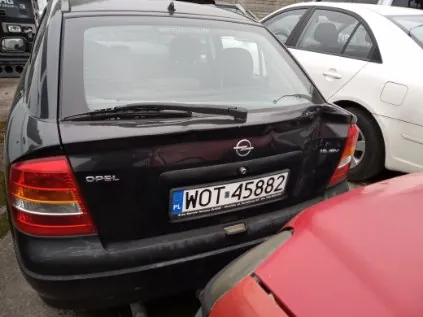 Czarny samochód marki Opel widziany od tyłu z tablicą rejestracyjną WOT-45882. Po prawej stronie widoczny biały samochód, a w dolnej części kadru czerwony pojazd. Tło stanowi parking z innymi samochodami.