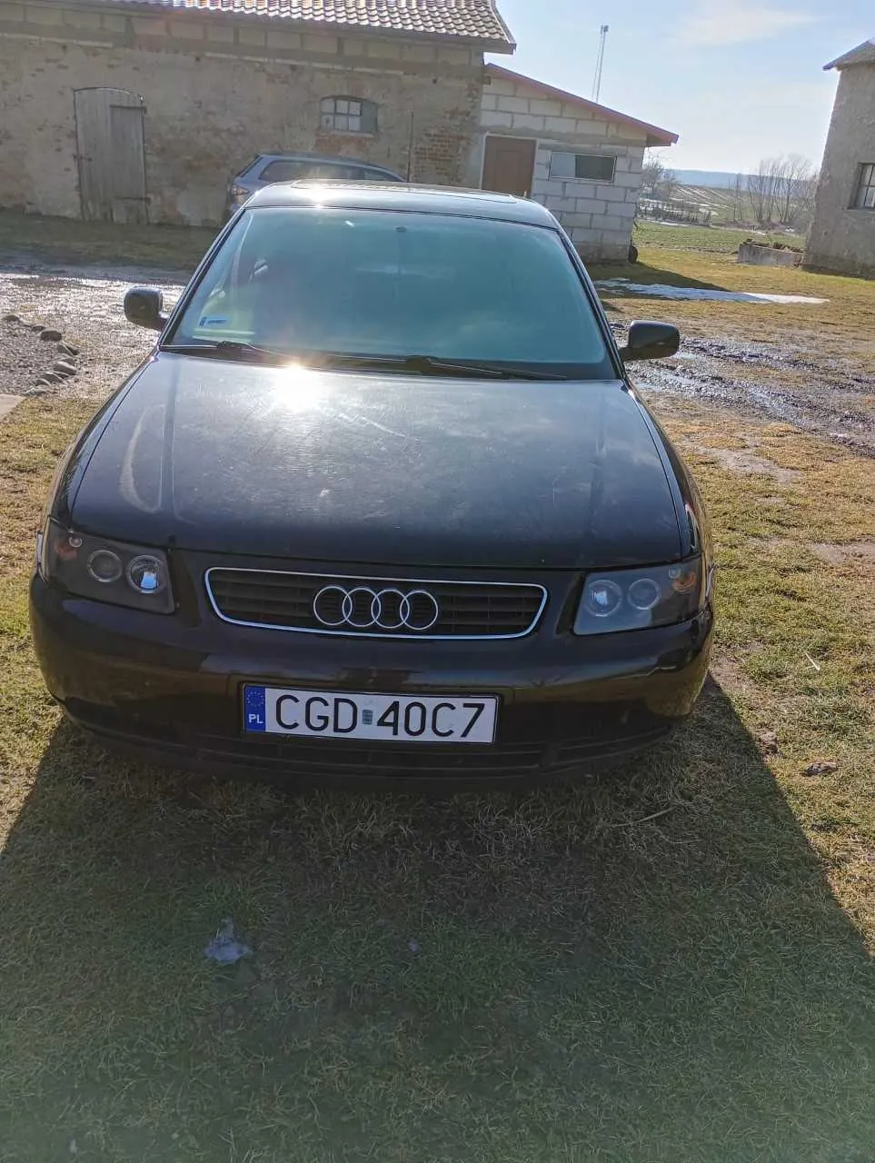 Czarny samochód marki Audi z przodu, z widocznym logo czterech pierścieni na atrapie chłodnicy. Tablica rejestracyjna PL CGD 40C7 umieszczona na przednim zderzaku. Samochód stoi na trawie, w tle stoją stare budynki z cegły i kamienia, częściowo zniszczone, z dachami krytymi dachówką. Na ziemi widoczne są kałuże i ślady błota. Słoneczny dzień, niebo jest jasne.