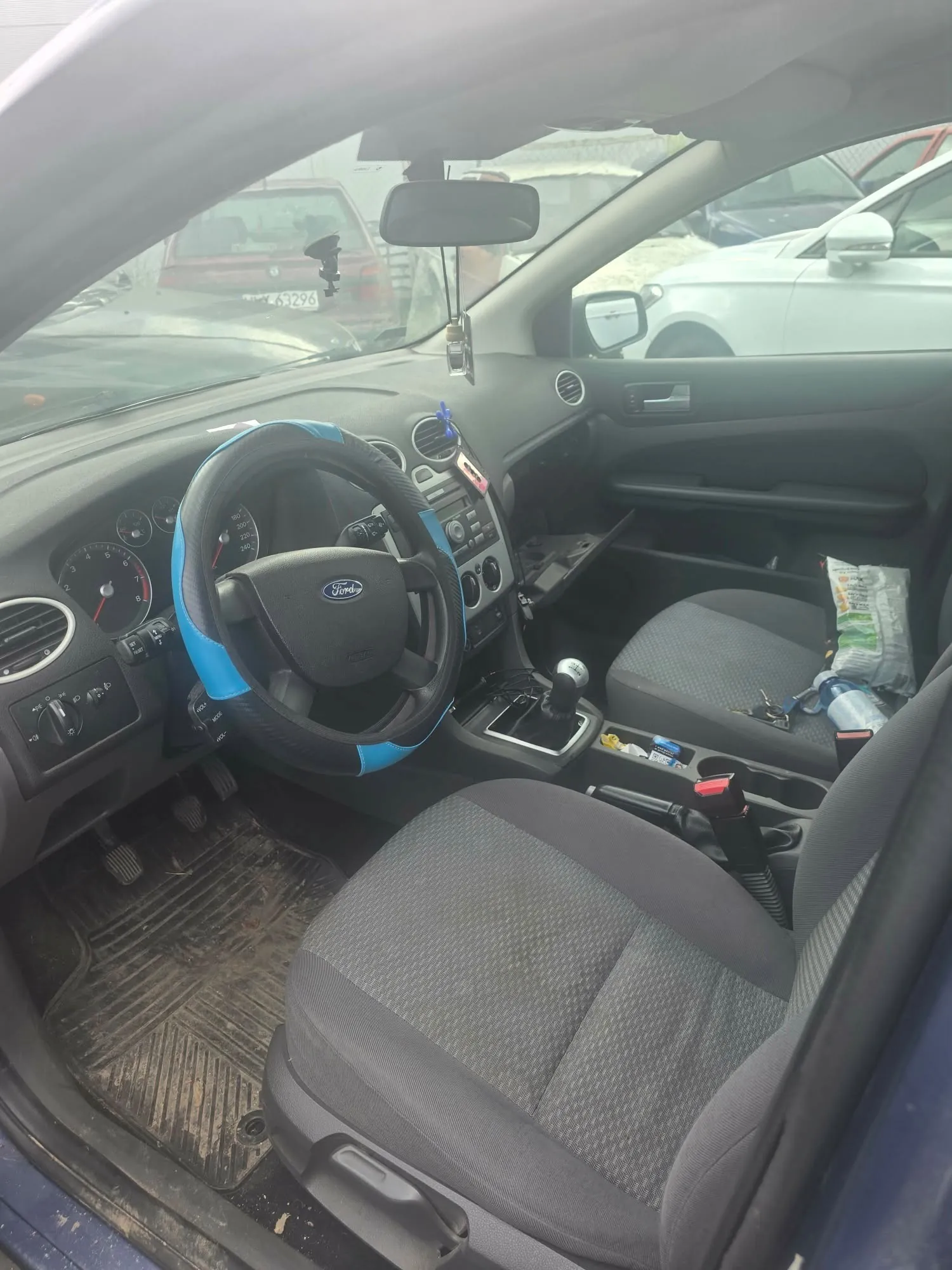Wnętrze samochodu marki Ford widziane z perspektywy otwartych drzwi kierowcy. Widoczny jest czarny fotel kierowcy i pasażera, kierownica z niebiesko-czarnym pokrowcem z logo Ford, deska rozdzielcza z zegarami oraz lewarek zmiany biegów. Na podłodze znajduje się gumowa mata. Przez przednią szybę widoczne są inne zaparkowane samochody.
