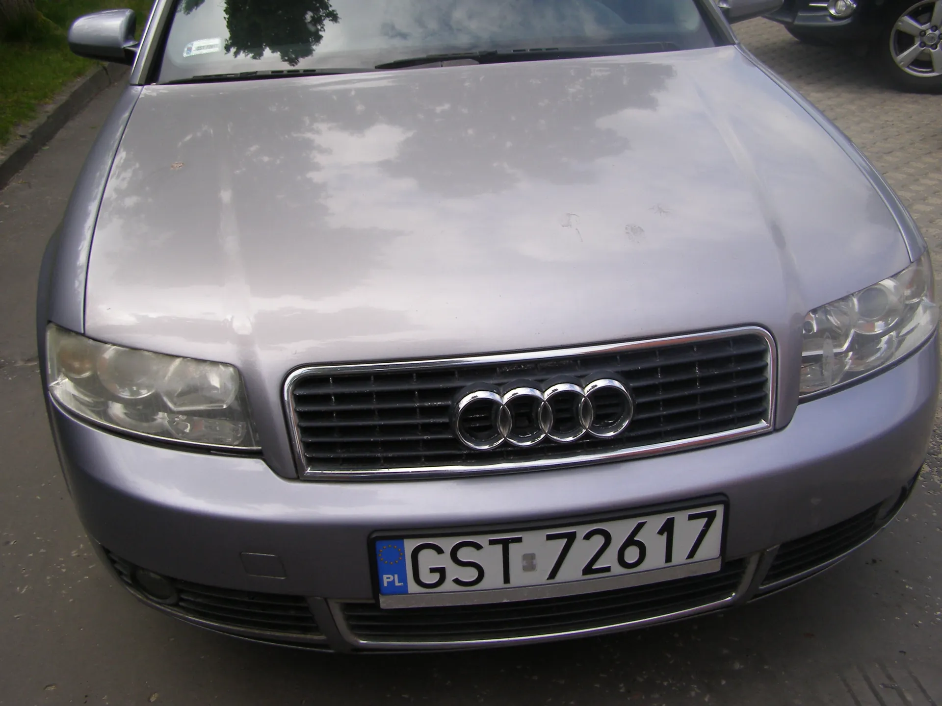 Przedni widok srebrnego samochodu marki Audi z widocznym czterokolcowym logo na atrapie chłodnicy. Na samochodzie umieszczona jest tablica rejestracyjna z białym tłem, czarnym napisem GST 72617 oraz niebieskim pasem z napisem PL po lewej stronie. W tle widoczne są fragmenty innego pojazdu, brukowana powierzchnia oraz zielona trawa po lewej stronie. Na masce odbijają się chmury i drzewa.