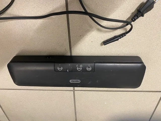 Czarny soundbar marki Morda leżący na jasnej podłodze z kafelkami. Na soundbarze widoczne są przyciski: zasilania, głośności, wejścia oraz logo marki. Obok urządzenia znajdują się czarne przewody, w tym wtyczka zasilająca i inny kabel zakończony złączem.