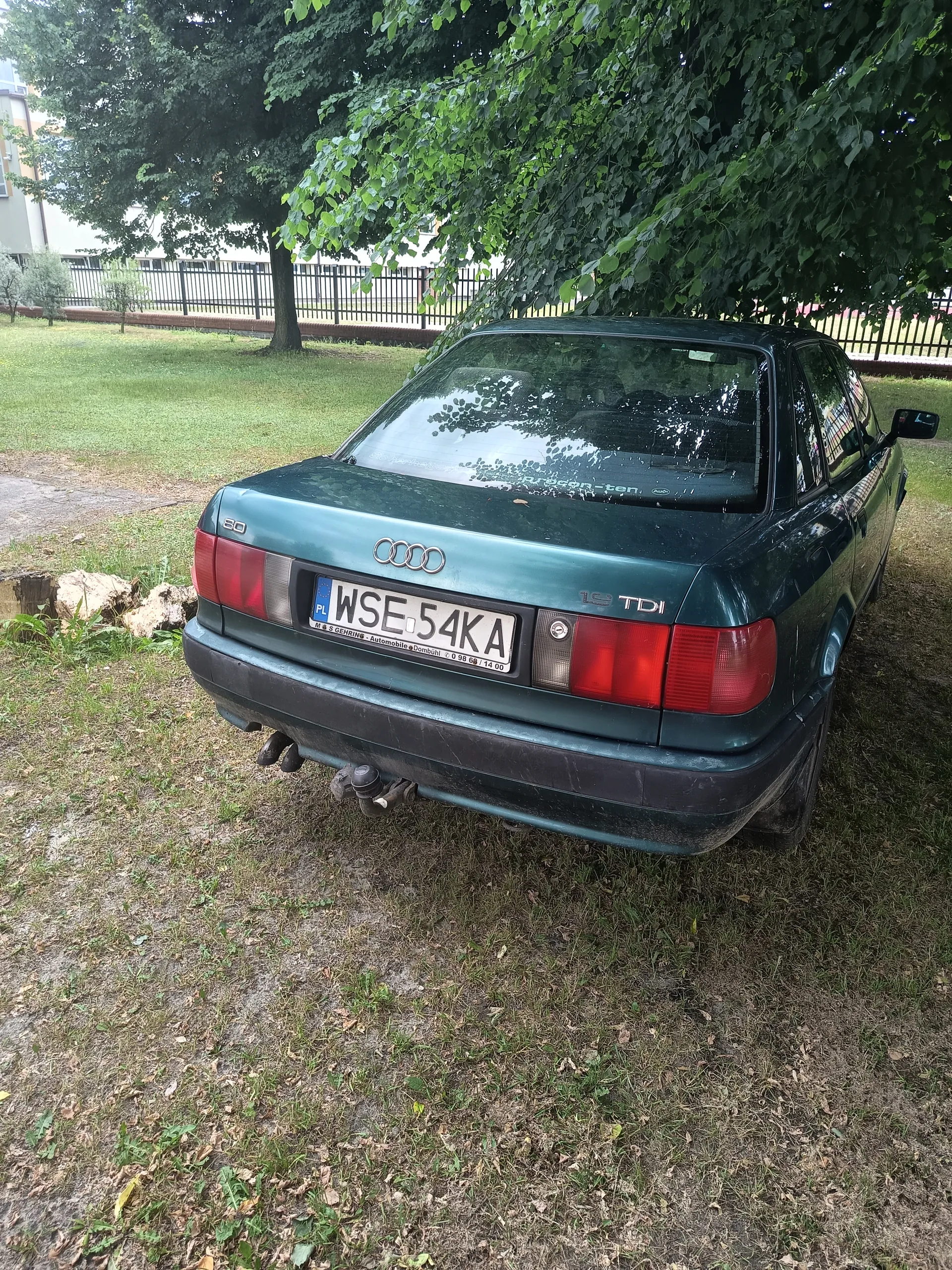 Zielony samochód Audi 80 TDI zaparkowany na trawie. Widoczny tył pojazdu z tablicą rejestracyjną WSE 54KA, emblematem Audi oraz napisem TDI. W tle drzewa, trawnik, czarny płot i budynek. Na szybie tylnej widoczny napis 'Propan-ten'.