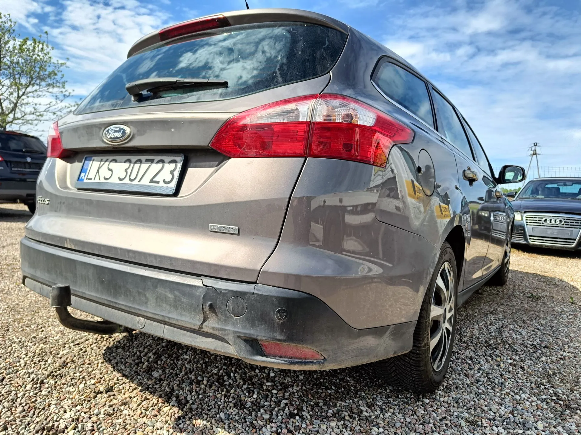Tylny widok brązowego samochodu marki Ford w nadwoziu kombi, zaparkowanego na żwirowym podłożu. Pojazd ma polską tablicę rejestracyjną o numerze LKS 30723. Na tylnym zderzaku widoczne jest pęknięcie oraz zamontowany hak holowniczy. W tle widoczne są inne samochody oraz niebo z chmurami.