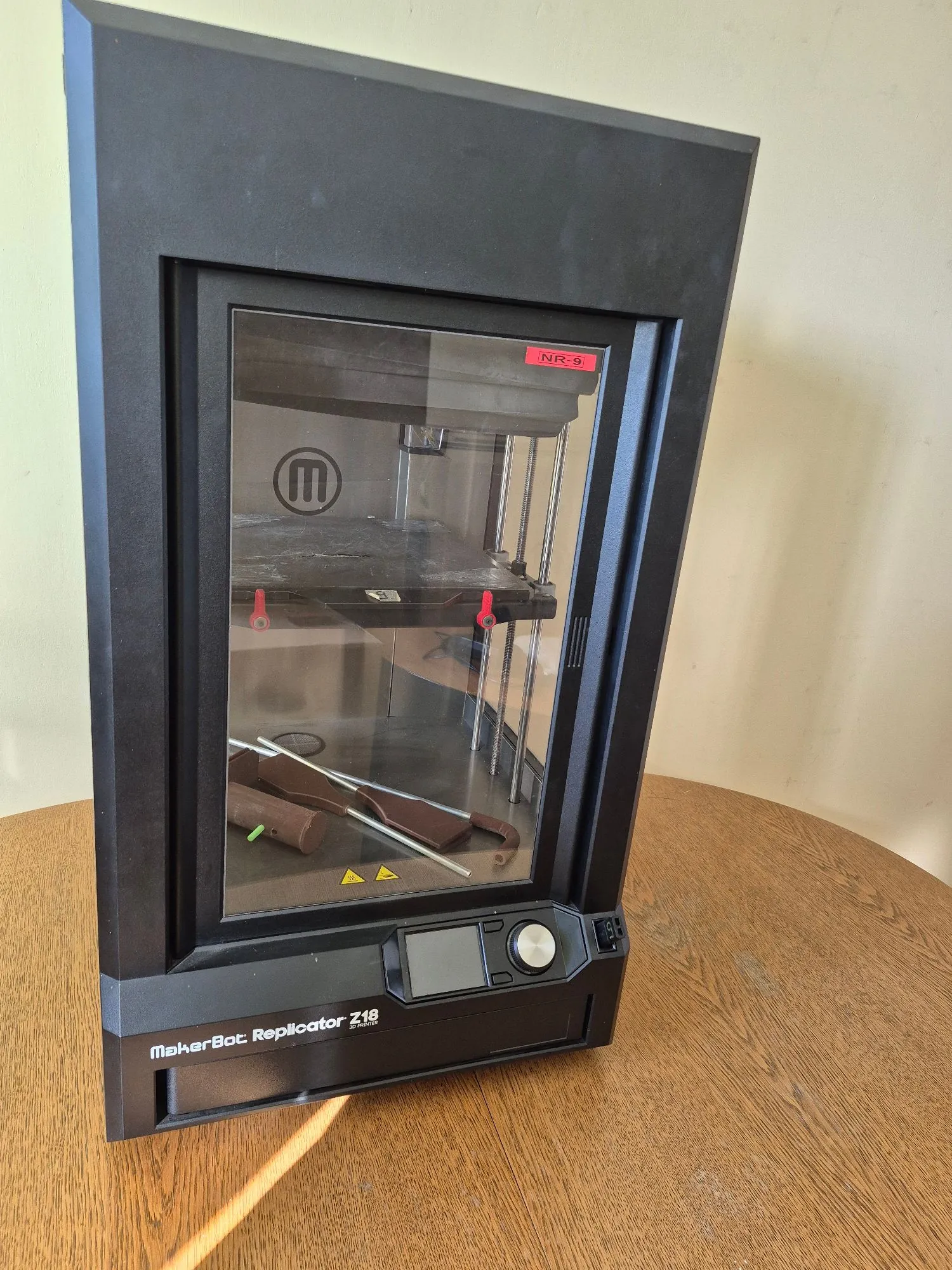 Czarna drukarka 3D marki MakerBot model Replicator Z18 z zamkniętą komorą roboczą. Wewnątrz urządzenia, na stole roboczym, znajdują się brązowe, wydrukowane elementy plastikowe przypominające części konstrukcyjne. Na dolnym panelu urządzenia widnieje napis 'MakerBot: Replicator Z18' oraz mały ekran LCD z pokrętłem sterującym. W górnej części komory znajduje się czerwona naklejka z napisem 'NR-9'.