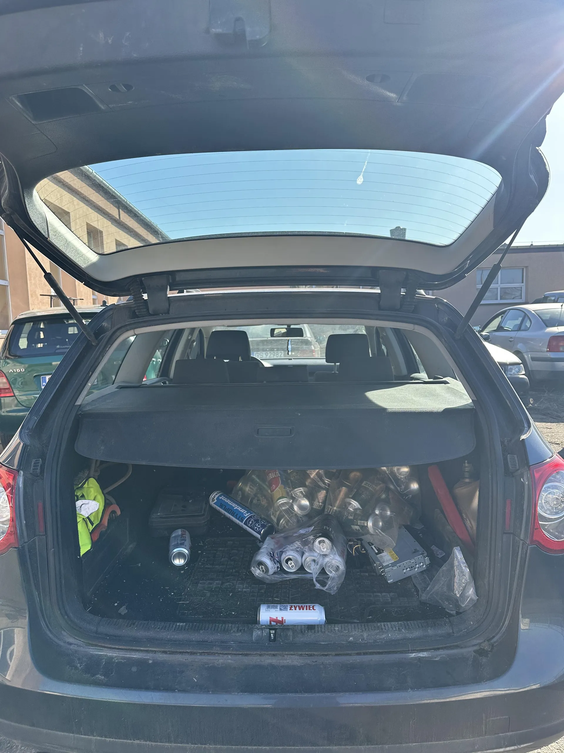 Otwarty bagażnik ciemnego samochodu z widocznymi przedmiotami: kilka puszek (w tym jedna z napisem ZYWIEC), opakowania z plastiku, zielony przedmiot po lewej stronie, czerwony przedmiot po prawej oraz elektroniczny element. W tle inne zaparkowane samochody i budynki. Słoneczna pogoda, niebo bezchmurne.
