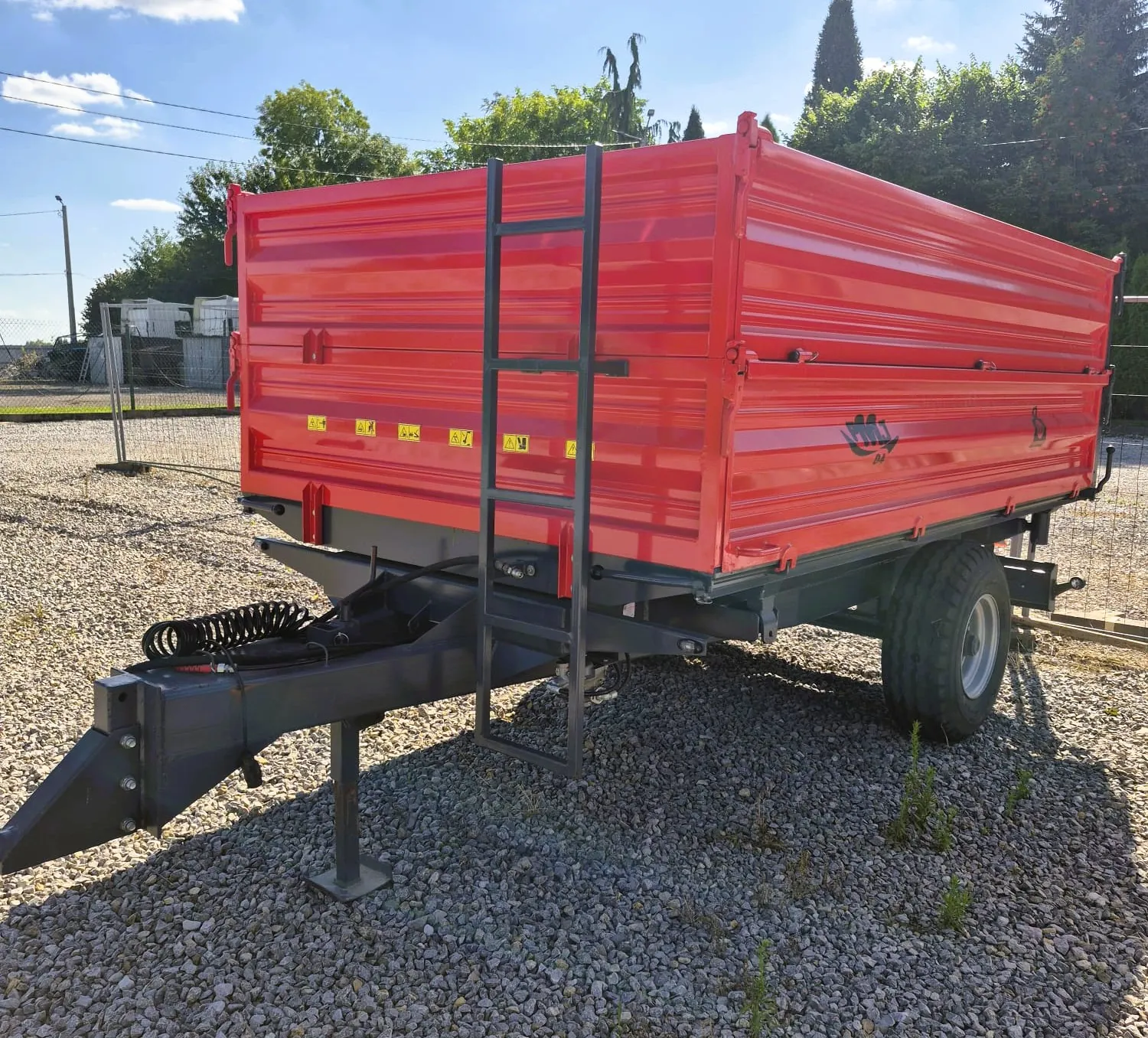 Czerwona przyczepa transportowa z wysokimi ścianami, zaparkowana na żwirowym podłożu. Przyczepa posiada czarną ramę, drabinę z boku oraz dyszel z przodu. Na bocznej ścianie widoczne są żółte tabliczki ostrzegawcze oraz logo producenta.