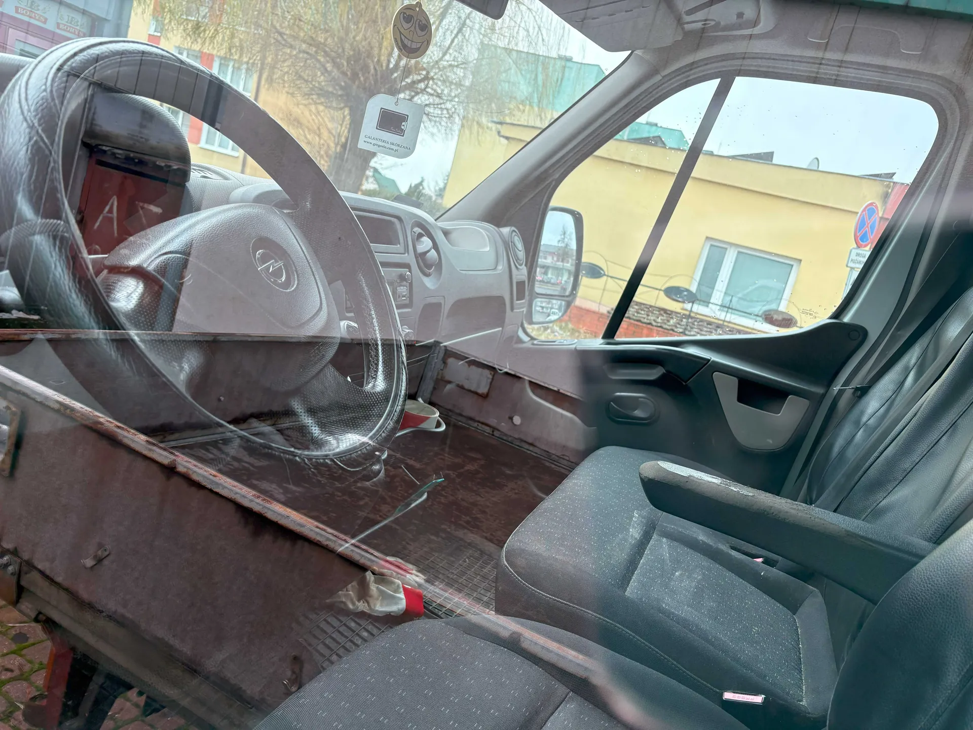 Wnętrze samochodu widziane z przodu, z kierownicą po lewej stronie. Widoczne są czarne siedzenia, deska rozdzielcza z przyciskami, lusterko boczne oraz przednią szybę. Na lusterku wiszą dwa przedmioty: żartobliwy uśmiechnięty znaczek i biały tag z napisem "GARANTOWANA JAKOŚĆ" oraz adresem www.garantowana.pl. Poza samochodem widoczne są żółte budynki, drzewo i znak zakazu postoju. Na podłodze wewnątrz samochodu leży czerwono-biały przedmiot oraz kubek.