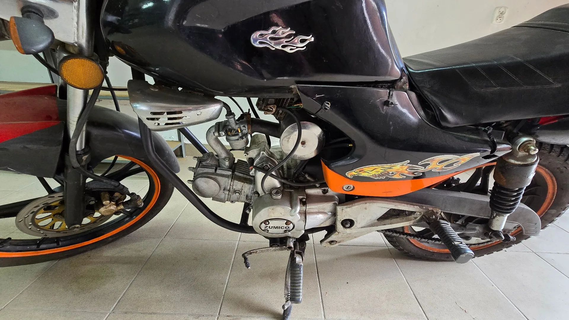 Zbliżenie na profil czarnego motocykla z pomarańczowymi akcentami na felgach i owiewkach. Na baku paliwa oraz bocznej owiewce widoczne są srebrno-żółte naklejki w kształcie płomieni. Na obudowie silnika znajduje się napis 'ZUMICO'. Motocykl stoi na podstopce w pomieszczeniu z jasną kafelkową podłogą.