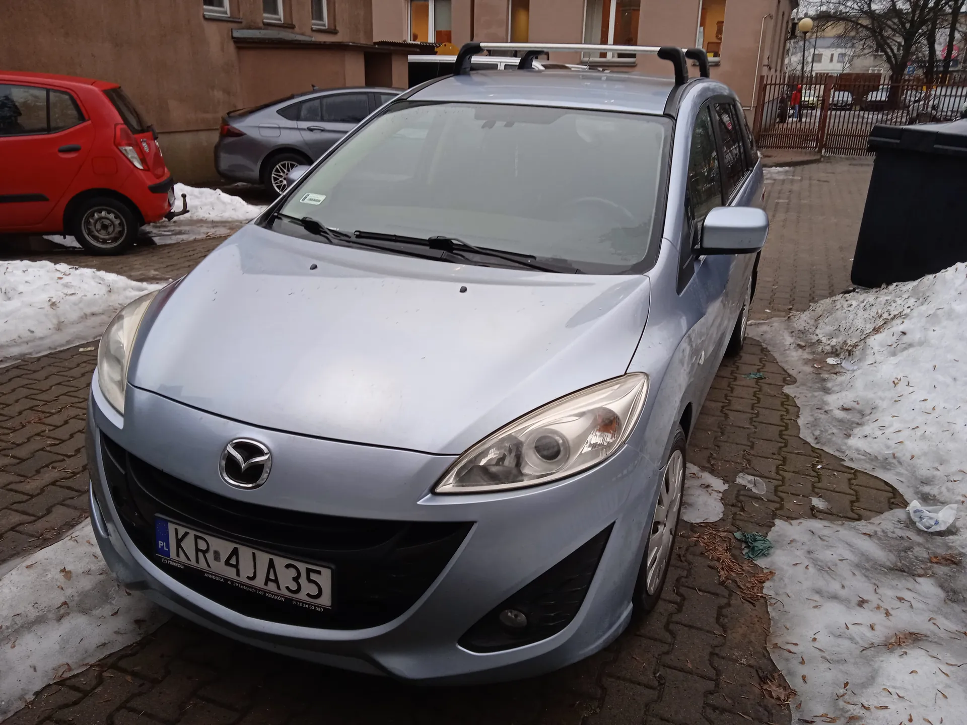 Srebrny samochód marki Mazda z tablicą rejestracyjną KR 4JA35 (PL) zaparkowany na brukowanej powierzchni. W tle czerwony i szary samochód, brązowy budynek z oknami, śnieg na ziemi oraz czarna pojemnik na śmieci. Na tablicy rejestracyjnej widoczny napis 'PL' oraz numer 'KR 4JA35'.