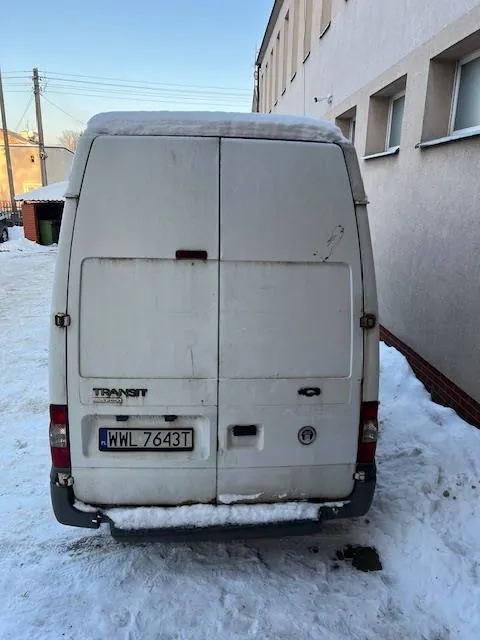 Tylny widok białego samochodu dostawczego marki Ford Transit zaparkowanego na śniegu. Pojazd ma dwie tylne klapy, tablicę rejestracyjną o numerze WWL 7643T oraz napis TRANSIT po lewej stronie. Na dachu samochodu znajduje się warstwa śniegu. W tle widoczny jest jasny budynek oraz zimowy krajobraz.