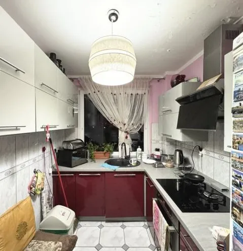 Kuchnia z białymi górnymi szafkami i bordowymi dolnymi szafkami. W centrum wisząca lampa z białym abażurem. Na prawo czarna płyta gazowa, po lewej zlewozmywak z kranem. Na blacie przed zlewem niebieska podkładka, obok czajnik i inne przedmioty. Okno z przezroczystą zasłoną, na parapecie doniczki z roślinami. Podłoga w czarno-białą mozaikę. Po lewej stronie miotła i kosz na śmieci. Na prawej ścianie kalendarz lub plakaty z małymi obrazkami. Ściany częściowo wyłożone jasnoszarymi płytkami.