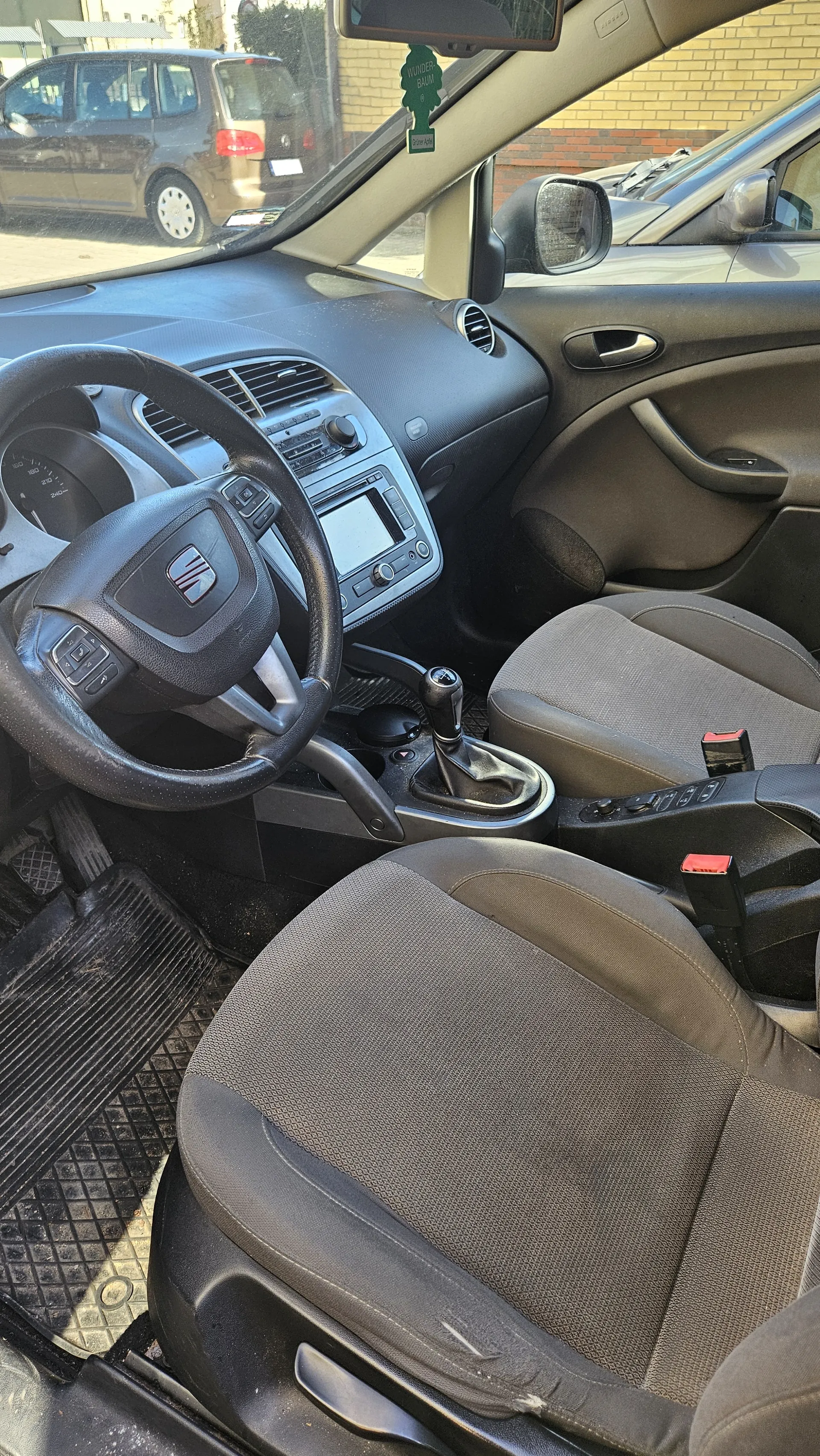 Wnętrze samochodu z widocznym kierownicą z logo, deska rozdzielcza z radioodtwarzaczem i klimatyzacją, dźwignią zmiany biegów, szarymi siedzeniami oraz dywanikami podłogowymi. Na lusterku wstecznym wisząca zielona ozdoba z napisem "WUNDER-BAUM Original". Przez przednią szybę widoczny zewnętrzny parking z innymi pojazdami, w tym brązowym samochodem osobowym.