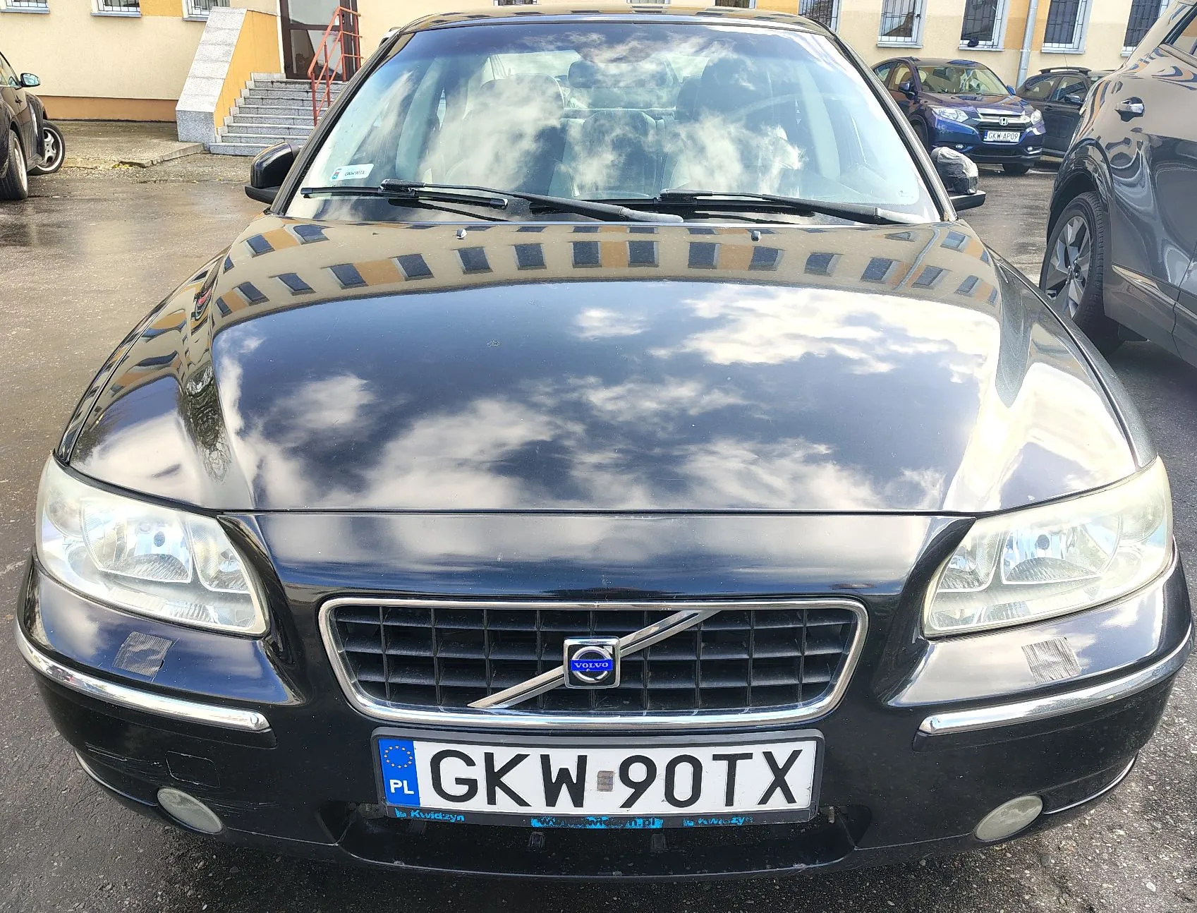 Widok z przodu na czarny samochód marki Volvo zaparkowany na mokrym placu. Na masce i przedniej szybie odbijają się chmury. Samochód posiada polską tablicę rejestracyjną o numerze GKW 90TX. W tle widoczne są inne zaparkowane samochody oraz żółty budynek.