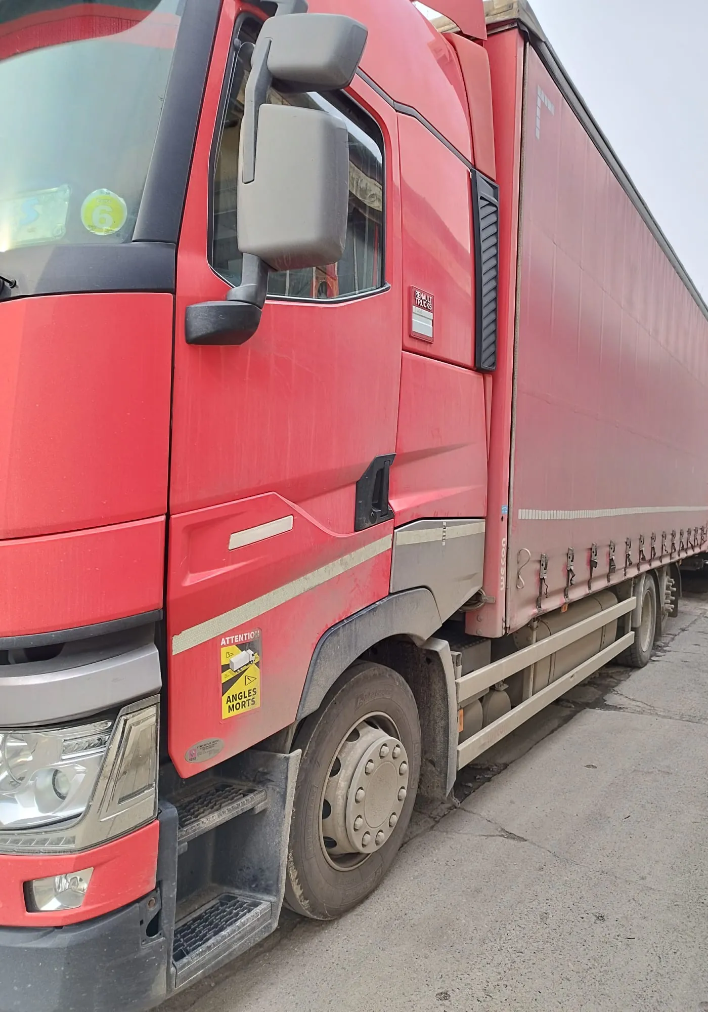 Czerwony ciężarówka z przyczepą zaparkowana na betonowej powierzchni. Kabina kierowcy jest czerwona z szarymi lusterkami bocznymi. Na przedniej szybie widoczny żółty okrągły naklejka z cyfrą 6. Na boku kabiny znajduje się żółta naklejka ostrzegawcza z napisem 'ATTENTION ANGLES MORTS' oraz czarno-żółtym symbolem. Przyczepa jest czerwona z białymi pionowymi liniami i metalowymi hakami. Widoczne są przednie reflektory, stopnie pod drzwiami kabiny oraz koła z metalowymi felgami. Tło stanowi szara betonowa powierzchnia i niebo.