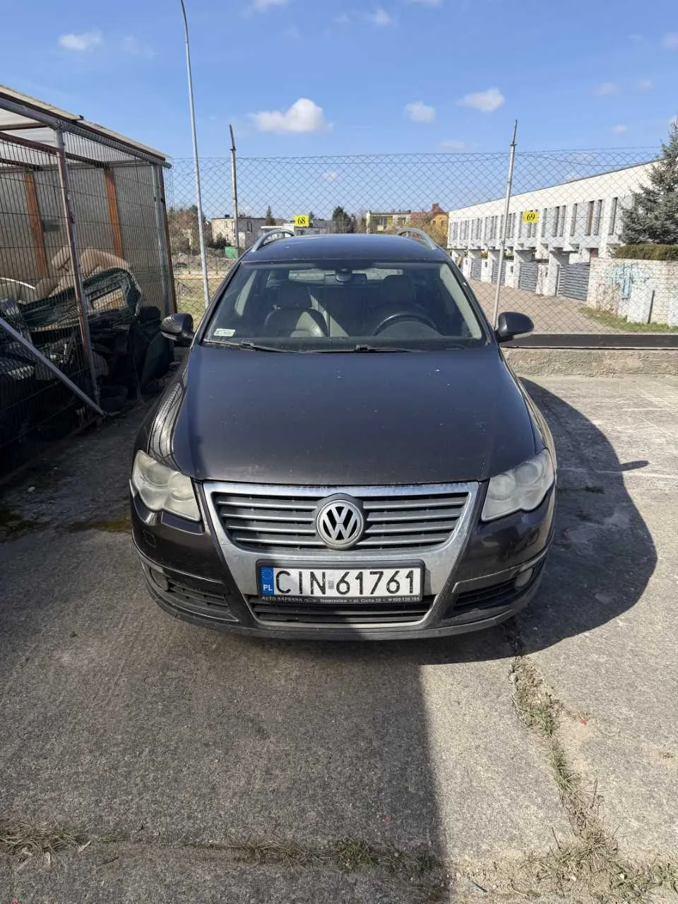 Czarny samochód marki Volkswagen z tablicą rejestracyjną PL CIN 61761 zaparkowany na betonowej powierzchni. W tle siatka ogrodzeniowa, po lewej stronie metalowa klatka z częściami samochodowymi, po prawej budynek z garażami i żółte tabliczki z numerami 68 i 69. Niebo jest błękitne z kilkoma chmurami.