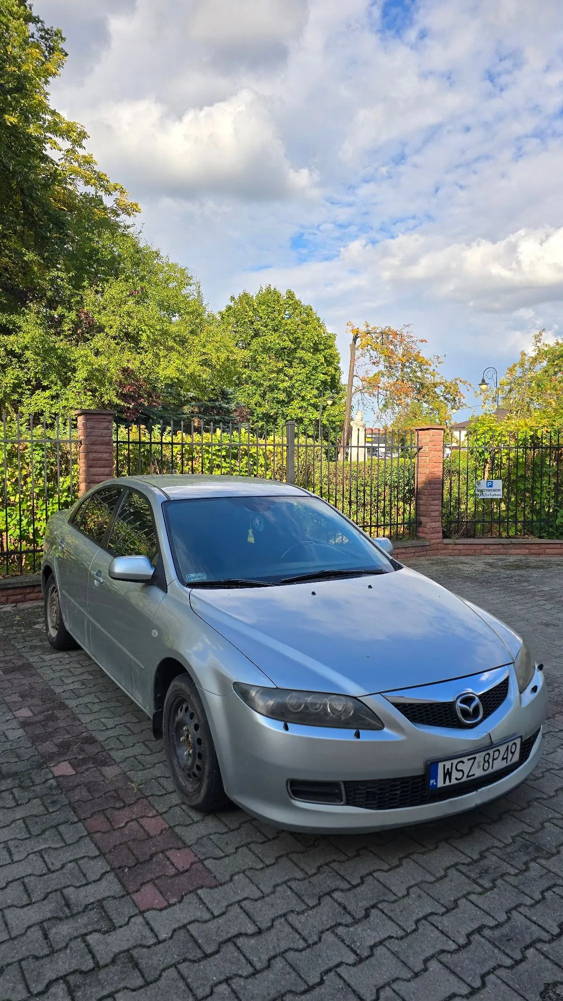 Srebrny samochód marki Mazda zaparkowany na kostce brukowej przed czarnym metalowym ogrodzeniem i zielonymi drzewami. Na samochodzie widoczna jest polska tablica rejestracyjna o numerze WSZ 8P49.
