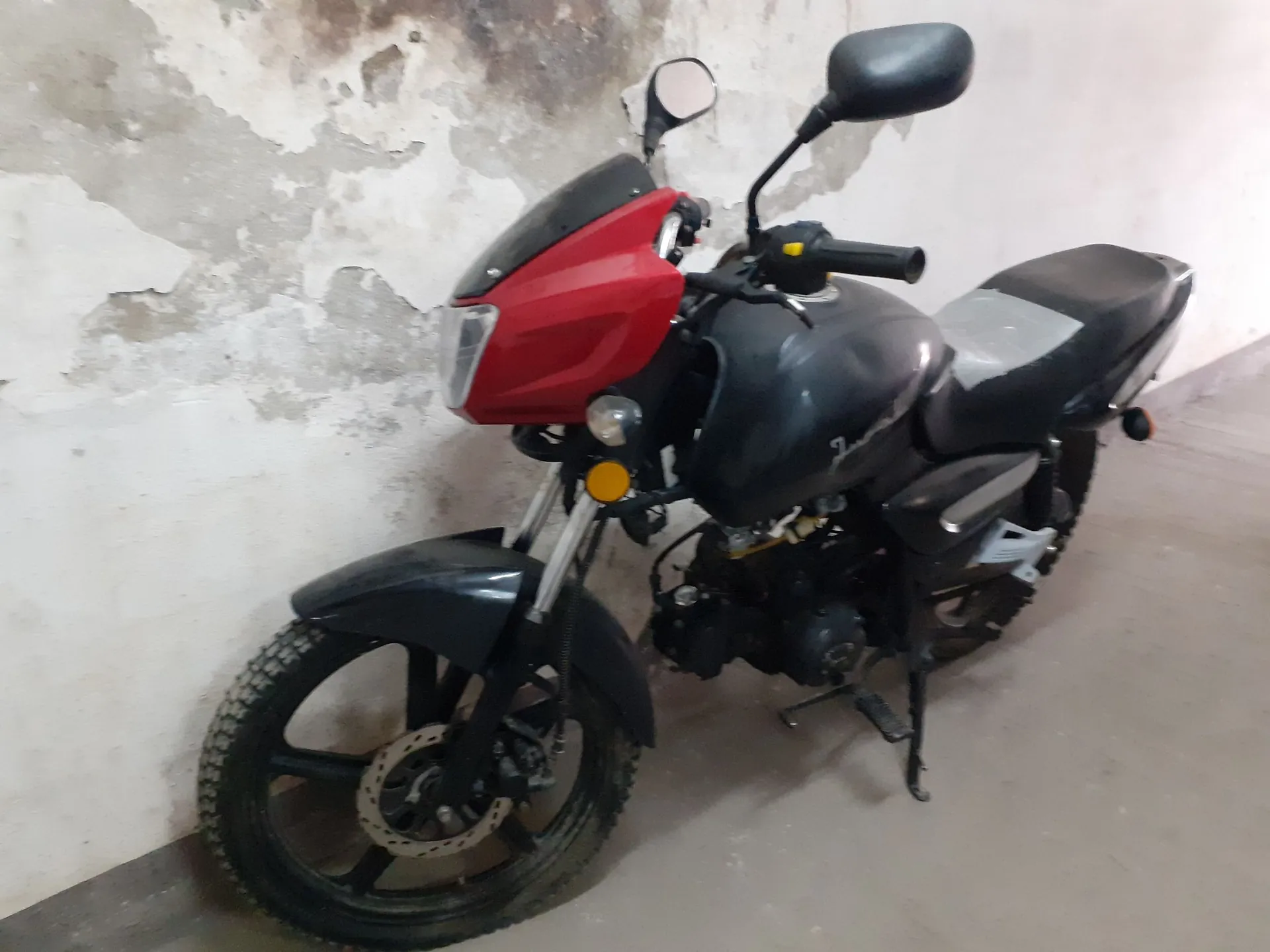 Czarny motocykl z czerwoną owiewką przednią, zaparkowany w pomieszczeniu na tle jasnej, zniszczonej ściany. Pojazd posiada dwa lusterka, czarne felgi z terenowymi oponami oraz widoczną konstrukcję silnika i podnóżek.