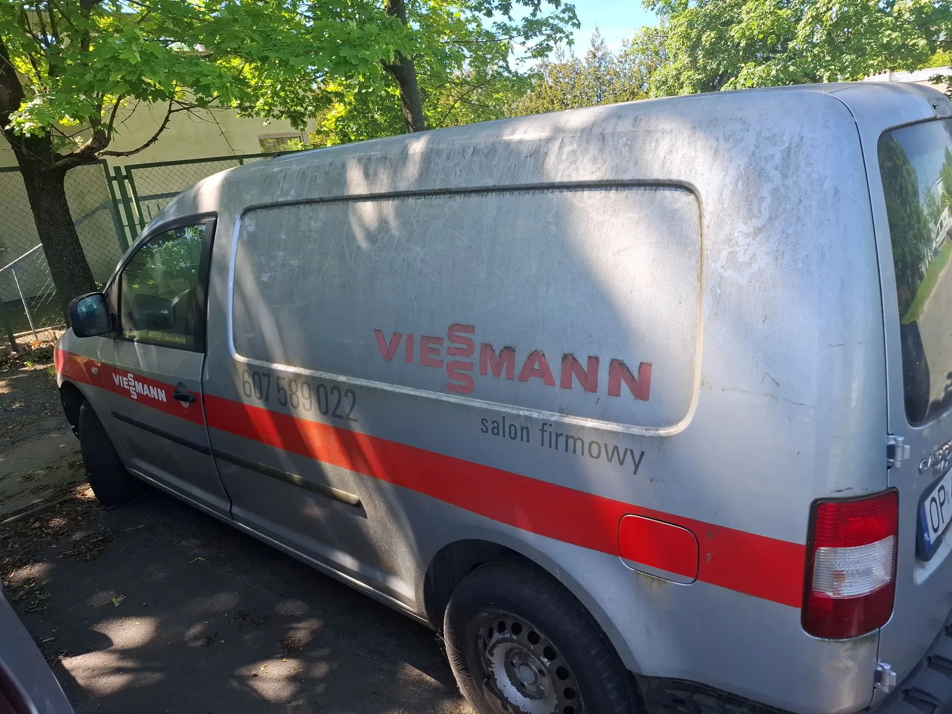 Srebrny furgon marki Volkswagen z czerwonym pasem u dołu nadwozia. Na boku pojazdu znajduje się czerwony napis 'VIESSMANN', numer telefonu '607 589 022' oraz mniejszy napis 'salon firmowy'. Samochód zaparkowany jest w cieniu drzew z zielonymi liśćmi.