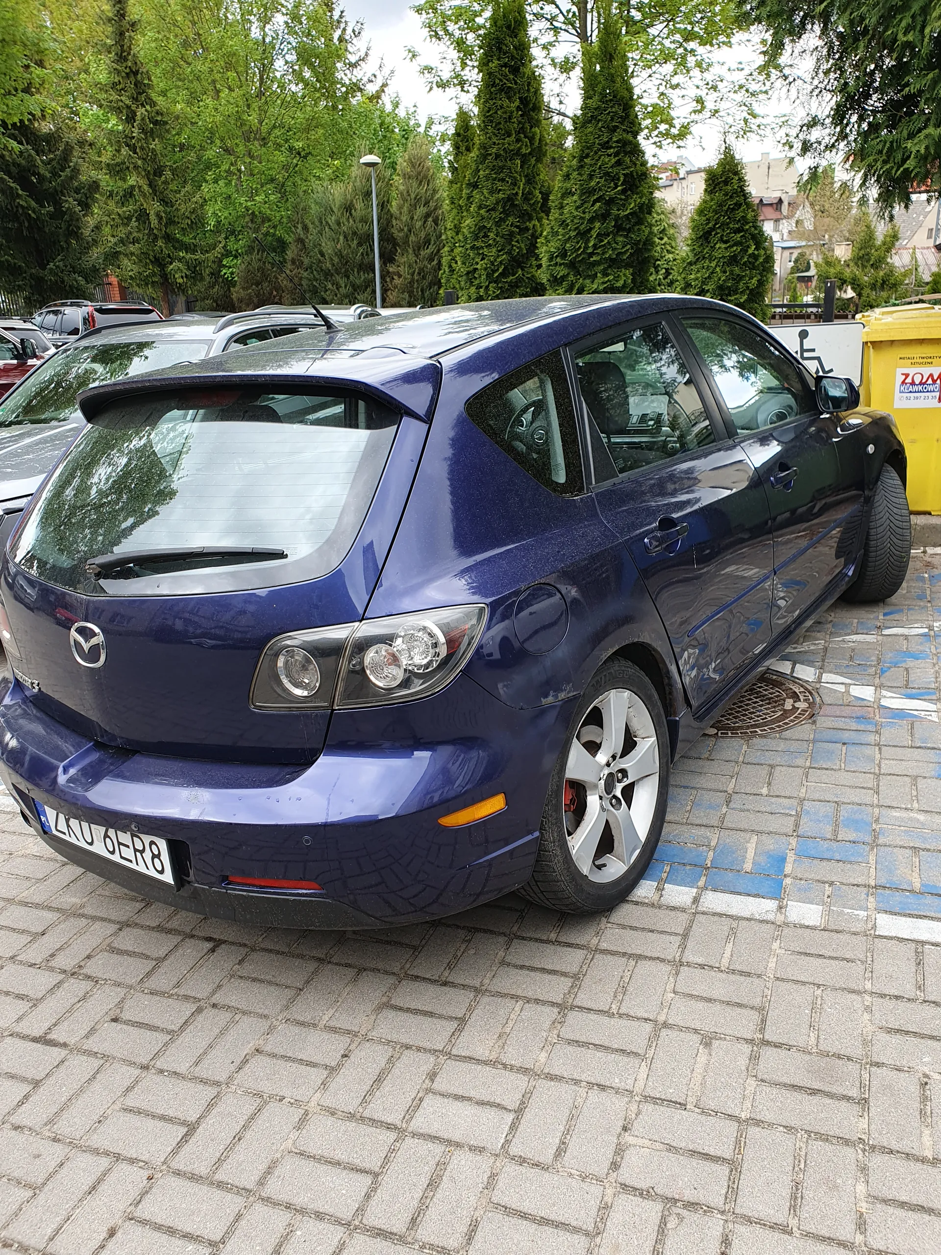 Ciemnoniebieski samochód marki Mazda zaparkowany na brukowanej powierzchni. Widoczna tablica rejestracyjna LKU 6E8. W tle żółty kontener z częściowo widocznym napisem ZOM, drzewa oraz inne zaparkowane pojazdy. Chodnik wyłożony płytkami z niebieskimi i białymi oznaczeniami.