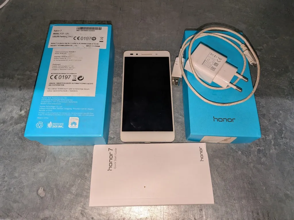 Zestaw produktów marki Honor ułożony na szarej powierzchni. W skład zestawu wchodzą: dwa niebieskie pudełka, biały smartfon Honor 7 z białymi ramkami, biała ładowarka sieciowa z podłączonym białym kablem USB oraz biała instrukcja obsługi z napisem 'honor 7 Quick Start Guide'. Na jednym z pudełek widoczne są etykiety z danymi technicznymi, kodem kreskowym oraz oznaczeniem CE 0197.