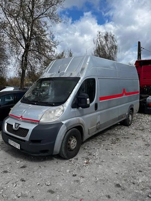 Srebrny furgon marki Peugeot z czerwoną linią biegnącą po boku, zaparkowany na żwirowym podłożu. Widoczny tabliczka rejestracyjna z napisem WY 1461V. W tle drzewa, częściowo zachmurzone niebo oraz czerwony pojazd. Furgon posiada ślady zużycia i zabrudzenia.