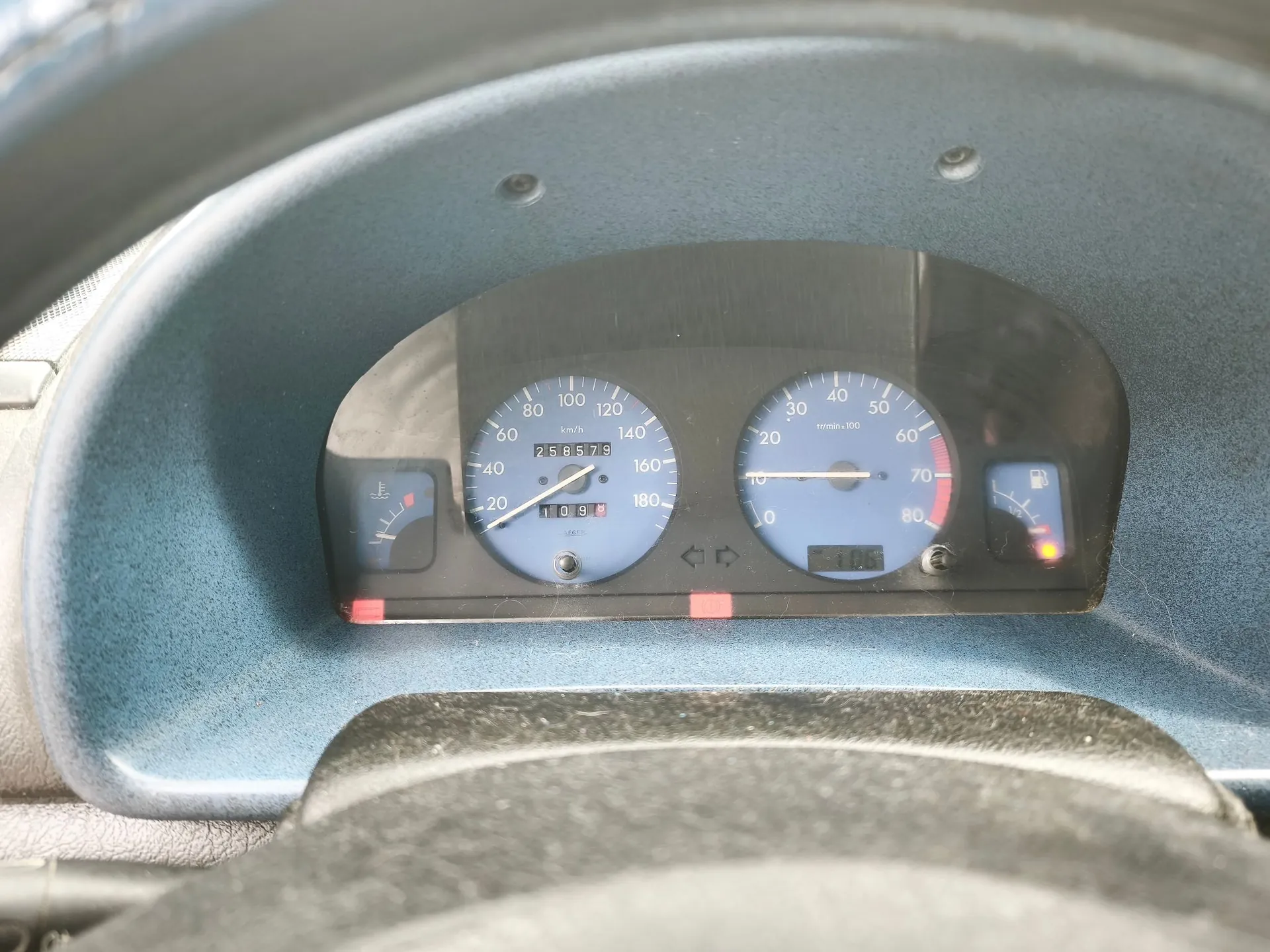 Zbliżenie na deskę rozdzielczą samochodu. Widoczny jest prędkościomierz z zaznaczoną skalą do 180 km/h, obrotomierz z zaznaczoną skalą do 80 x 100 obr./min oraz wskaźniki poziomu paliwa i temperatury silnika. Pomiędzy głównymi zegarami znajduje się licznik przebiegu. Całość osadzona jest w niebieskiej obudowie kokpitu.