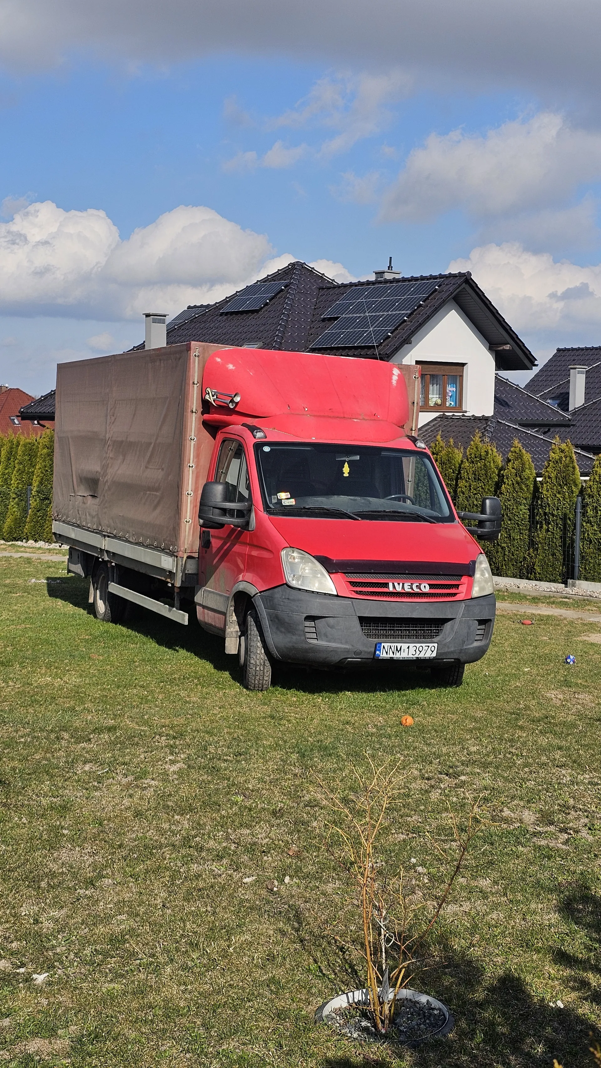 Czerwony samochód dostawczy marki IVECO z brązowym przyczepem zaparkowany na trawniku. Widoczna tablica rejestracyjna NNM 13979. W tle dom z czarnym dachem i panelami słonecznymi, zielone krzewy, niebo z białymi chmurami. Na trawniku znajduje się młode drzewko bez liści, niebieski przedmiot oraz pomarańczowa piłka. Trawa częściowo wysuszona.