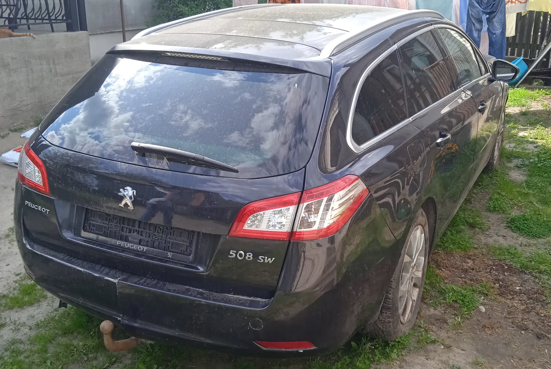 Czarny samochód Peugeot 508 SW widziany od tyłu, zaparkowany na trawie i ziemi. Widoczne logo Peugeot z lwem, napisy 'PEUGEOT' po lewej stronie bagażnika oraz '508 SW' po prawej. Brudna karoseria, reflektory tylne, szyba tylnej klapki z odbiciem nieba i chmur. W tle betonowa ściana, pranie wiszące na sznurze oraz ogrodzenie.