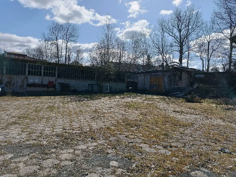 Zdjęcie przedstawiające zniszczone, opuszczone budynki przemysłowe z dużymi oknami i zniszczoną elewacją. Przed budynkami znajduje się rozległy, zniszczony plac z betonowych płyt, przez które przerasta trawa i mchy. W tle widoczne są bezlistne drzewa na tle niebieskiego nieba z białymi chmurami.