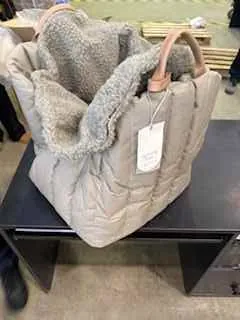 Beżowa pikowana kurtka z kapturem wykończonym futerkiem, z białą etykietą przy sznurku. Kurtka leży na czarnym stole. W tle widoczne są drewniane deski i betonowa podłoga. W prawym dolnym rogu obrazu widać fragment stopy w czarnej obuwie.