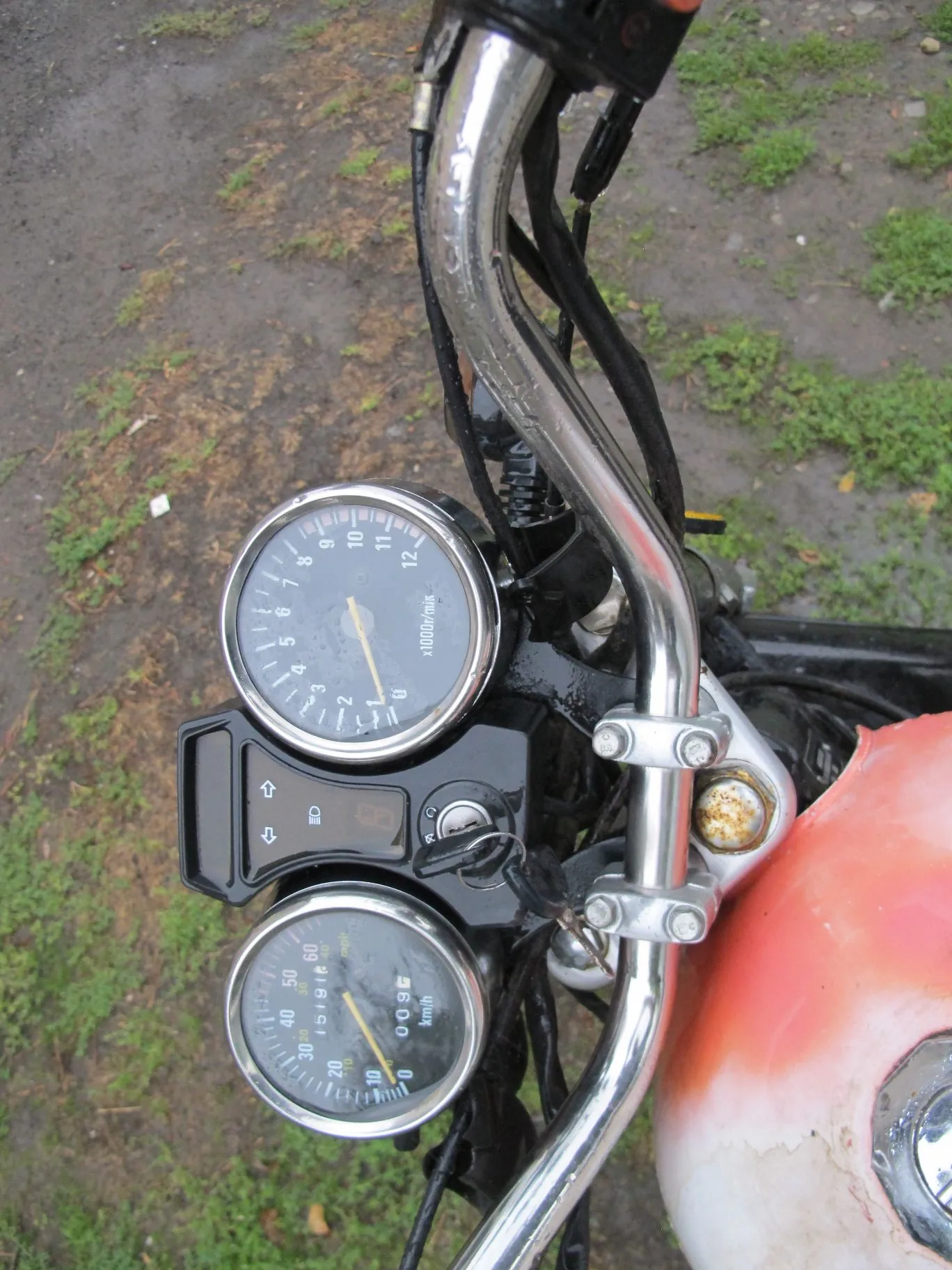 Zbliżenie na kierownicę i zestaw wskaźników motocykla. Widoczne są dwa okrągłe zegary: górny to obrotomierz z oznaczeniem 'x1000r/min', a dolny to prędkościomierz z oznaczeniem 'km/h'. Pomiędzy zegarami znajduje się czarny panel z przełącznikami i stacyjką z włożonym kluczykiem. Po prawej stronie widoczny jest fragment czerwono-białego baku paliwa oraz chromowana kierownica.