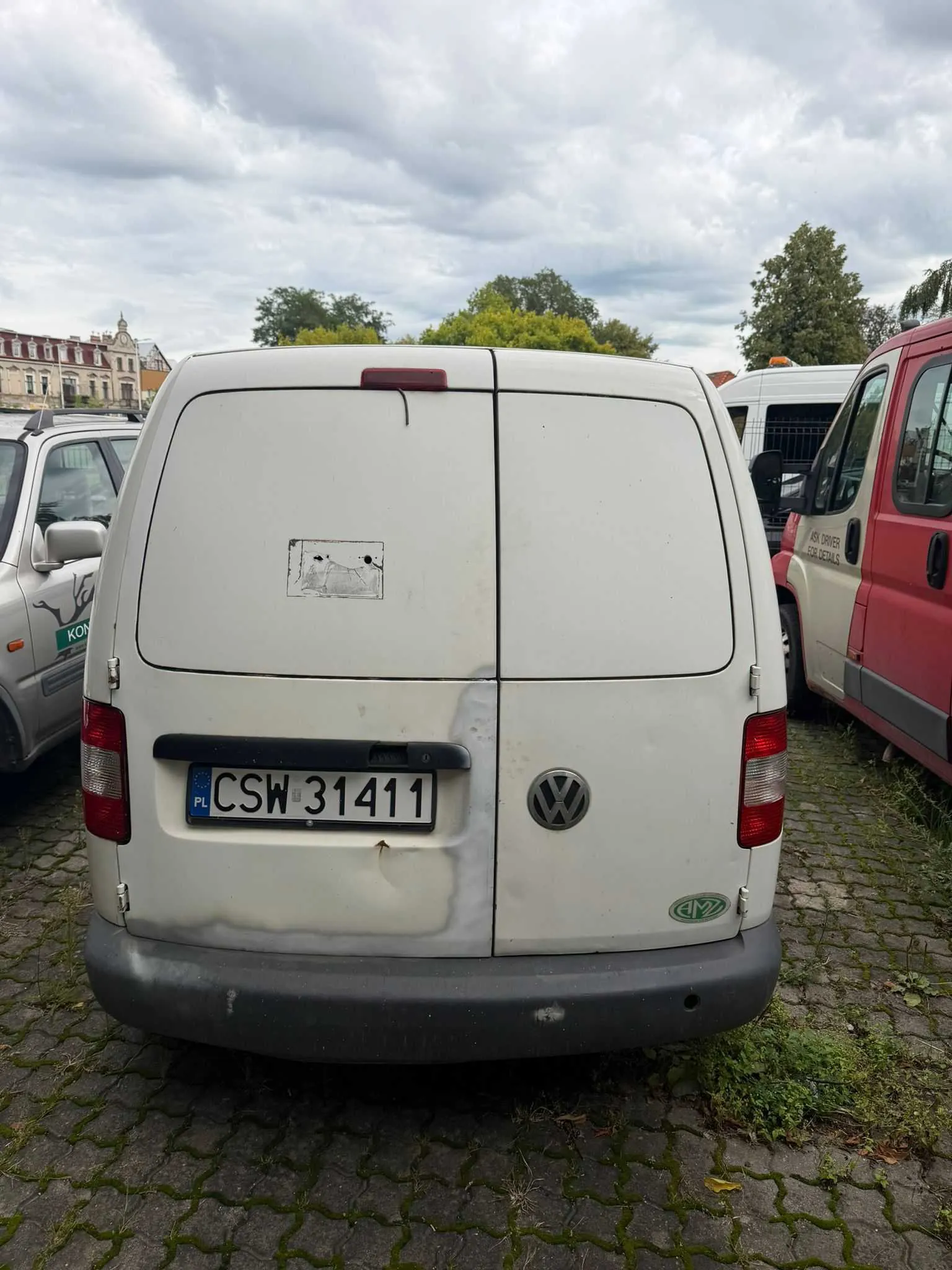 Tylny widok białego vana marki Volkswagen zaparkowanego na kostce brukowej. Pojazd ma tablicę rejestracyjną o numerze CSW 31411. Na tylnych drzwiach widoczne są ślady naprawy blacharskiej i malowania w kolorze szarym. Po lewej stronie stoi inny biały samochód, a po prawej czerwony van.