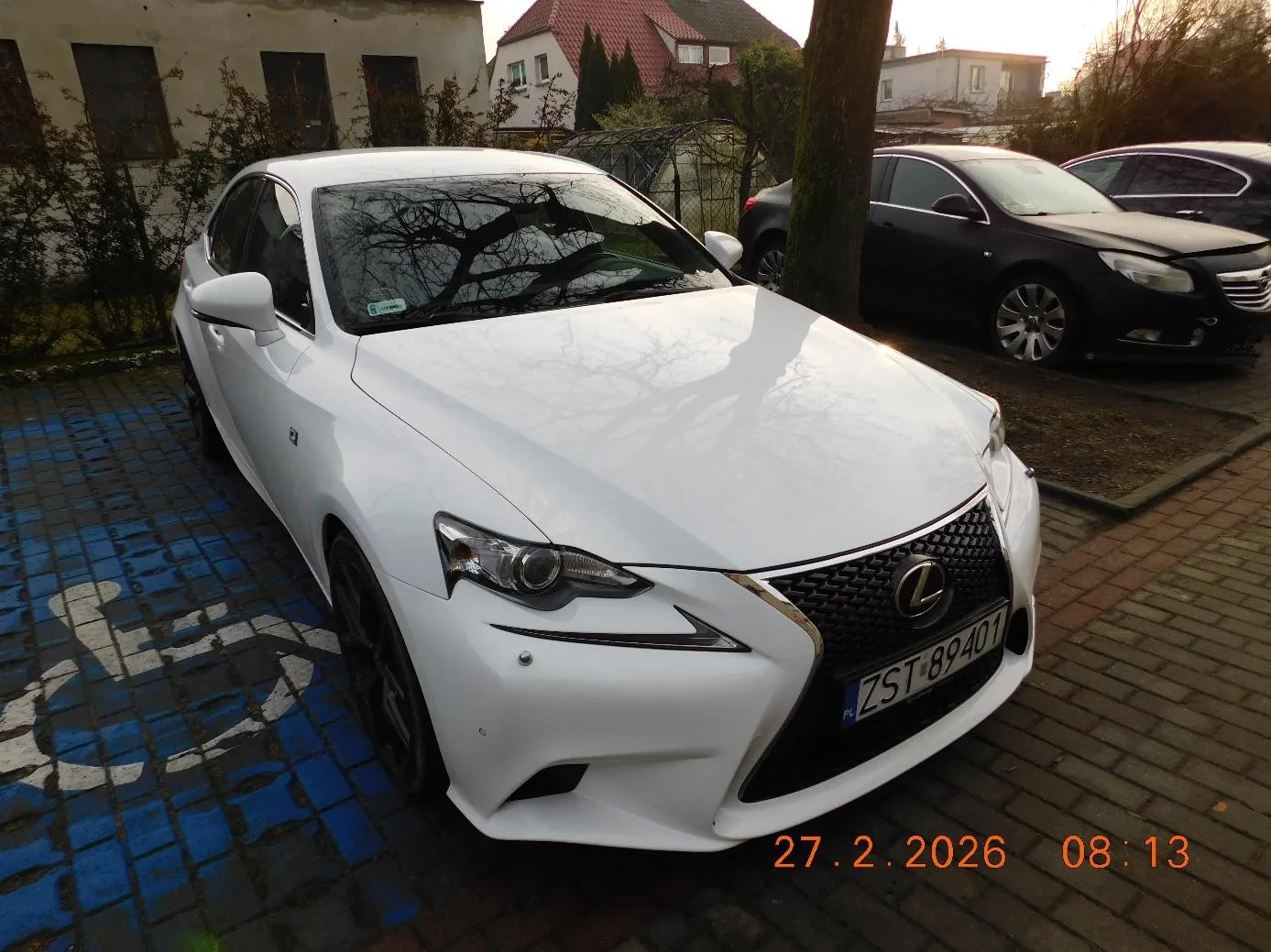 Biały samochód marki Lexus z tablicą rejestracyjną ZST 89401 zaparkowany na niebiesko-białym miejscu dla rowerów. W tle czarny samochód, drzewo, budynki z czerwonymi dachami oraz krzewy. Na dole zdjęcia widoczny pomarańczowy znaczek z datą 27.2.2026 i godziną 08:13.